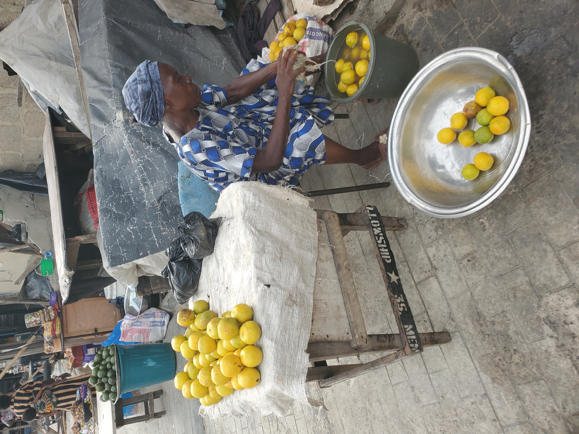 This media file depicts an outdoor market scene in Lagos, Nigeria, centered around an elderly vendor. The main subject, an older person likely a woman, is seated, wearing a blue and white patterned long-sleeved top and a blue head covering. She is actively engaged with a fibrous material, possibly processing or cleaning the fruits she sells.
The stall primarily features large, round yellow-green fruits, resembling oranges, grapefruits, or pomelos. These fruits are displayed in various ways: a large metal basin contains several, some in varying stages of ripeness; a green bucket filled with water also holds many, suggesting they might be kept cool or cleaned; and a substantial pile rests on a white cloth draped over a simple wooden stand. Another green plastic bucket, further to the left, is filled to the brim with avocados.
The setting is characteristic of a bustling market environment. Structures are informal, with black and light-colored tarpaulins providing shade, supported by wooden beams. The ground is paved with concrete blocks. In the background, other market activities and stalls can be faintly seen. The scene is brightly lit by daylight.
Visible text includes "DANGOTE" on a white mesh bag near the vendor's left, "DANGOTE CEMENT" on a blue sack further back, and "LOXSHIP", "35.", and "MES" (possibly part of "MESSENGER") printed on a wooden beam of the stall. The activity suggests a vendor preparing or selling produce in a lively market setting.