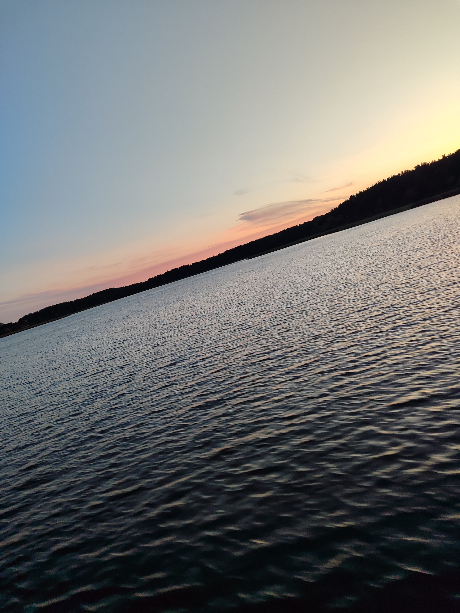 A low-angle, wide shot shows a body of water with a treeline in the distance at sunset. The water occupies the foreground and midground, with gentle ripples reflecting the sky's colors. The treeline, a dark silhouette against the sky, forms the horizon. The sky above the treeline transitions from a pale blue to a soft pink and then to a warm yellow near the horizon, indicating the sun is setting. The image is tilted, giving a dynamic perspective to the scene. There are no people or identifiable objects on the water or the shore.