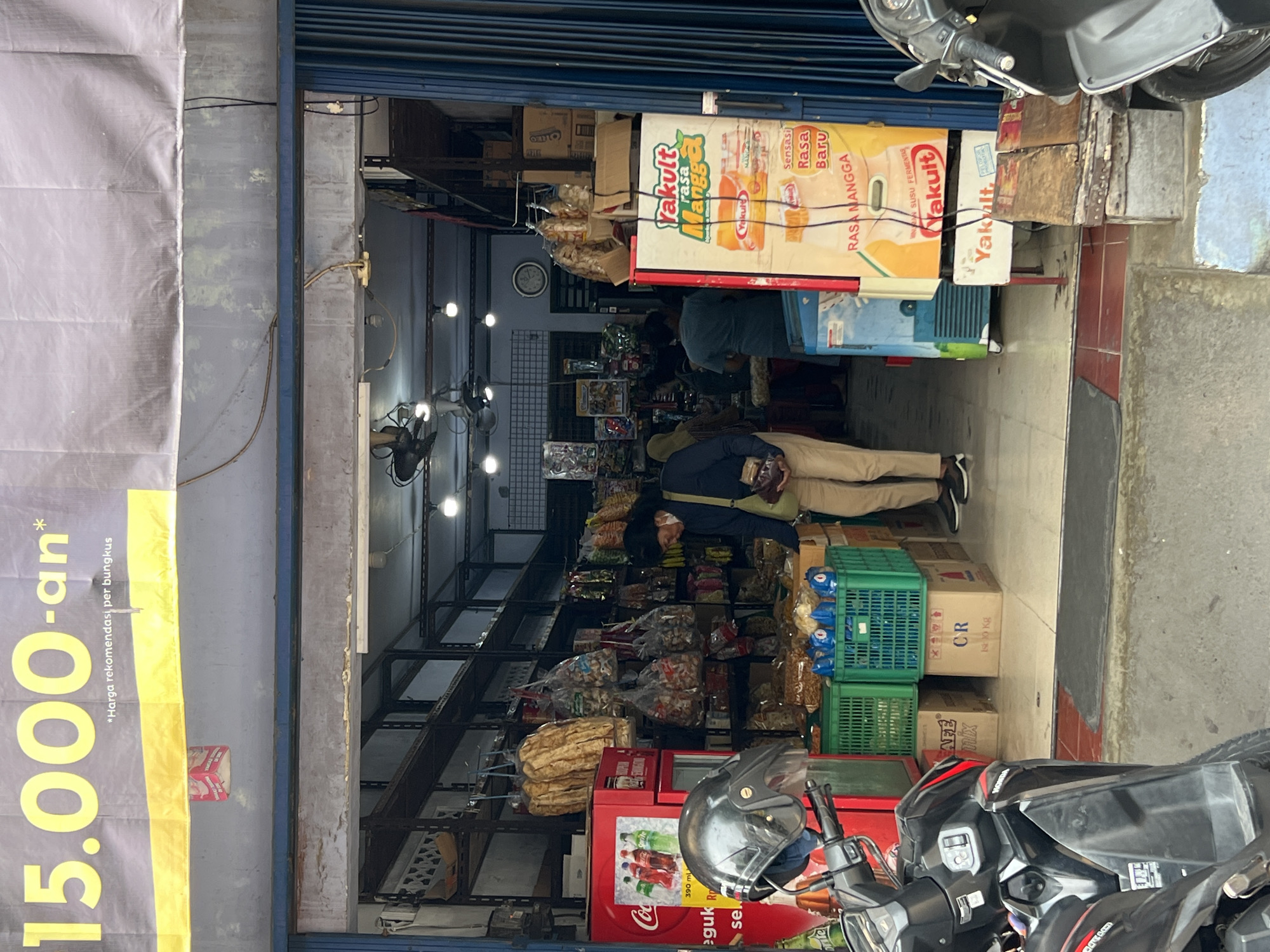 This is an exterior medium shot of a small storefront in Yogyakarta, Indonesia. A large banner with yellow lettering that reads "15.000-an*" is displayed prominently above the entrance. Below this banner, a red Coca-Cola refrigerator is partially visible on the left, and a Yakult refrigerator is on the right. Inside the store, shelves are stocked with various packaged goods, including snacks and drinks. Several people are inside the store, with one person in the foreground wearing a blue shirt and beige pants, carrying a dark bag over their shoulder. Two motorcycles are parked in front of the store, one on the left and one on the right, partially obstructing the view of the storefront. The ground in front of the store is paved with concrete and red bricks. The store is likely a small convenience store or a roadside minimarket.
