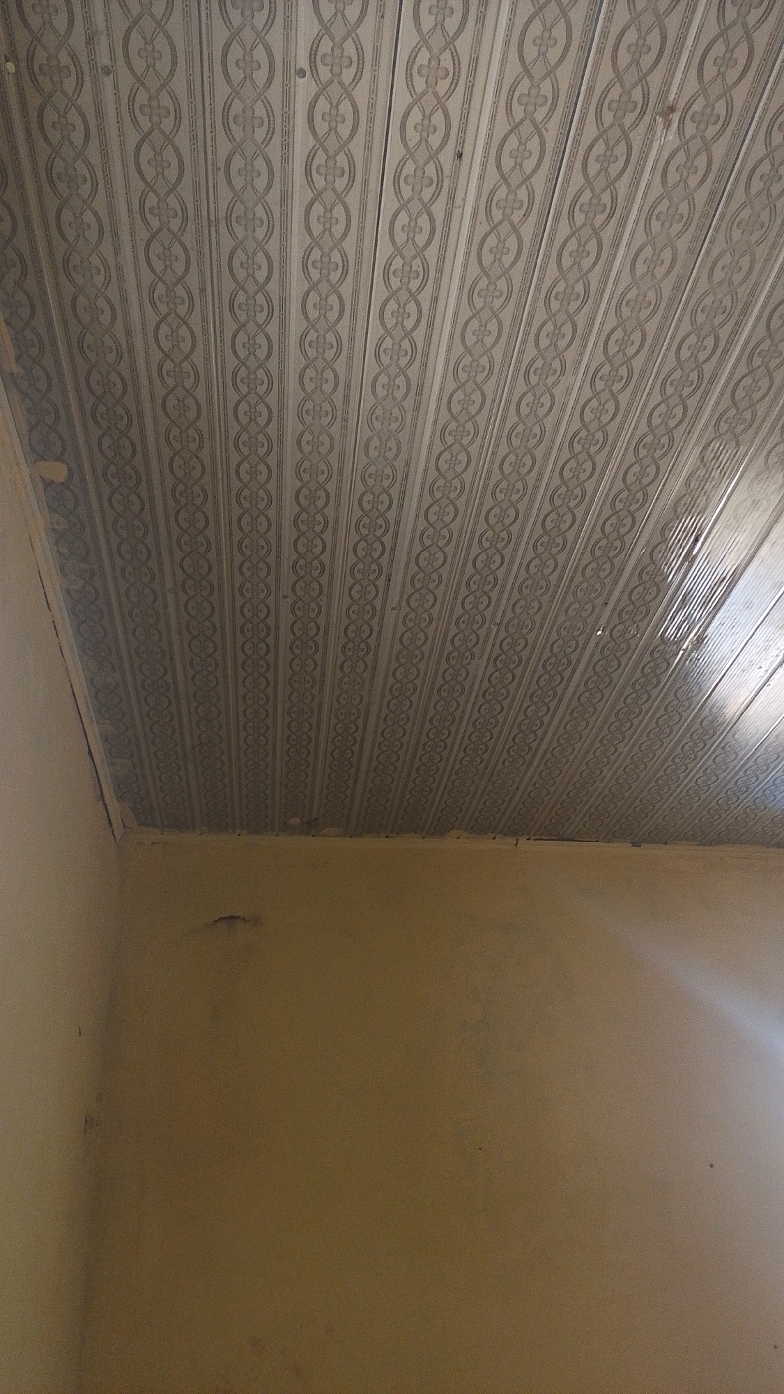The image shows the interior of a room with a decorated ceiling. The ceiling is made of light-colored panels with a repeating pattern of interlocking oval shapes and floral elements. The walls are a uniform, muted yellow color and appear to be a solid material, possibly plastered or painted. There is a corner visible where the ceiling meets the walls. The lighting suggests an indoor environment, with some areas of the ceiling appearing brighter, possibly due to light sources not directly visible in the frame. The overall impression is that of a simple, finished room. This image was taken in Balewa, Nigeria.