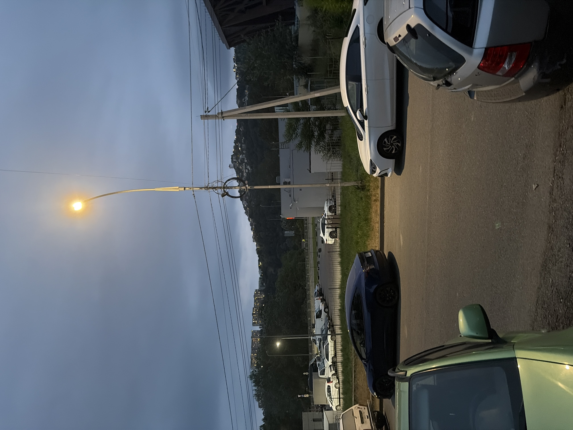 The image depicts a street scene in Sochi, Russia, during twilight. A street lamp illuminates the dusk sky, casting a warm glow on the surrounding environment. Several vehicles are parked along the roadside, including a dark blue Tesla Model 3, a white sedan, and a silver hatchback. In the background, a tree-lined hill rises, dotted with buildings that glow with interior lights, suggesting a residential area. The sky is overcast, contributing to the subdued lighting. A few power lines crisscross the frame, indicative of urban infrastructure. The foreground features the partial view of a light green car, its side mirror visible. The overall atmosphere is tranquil and urban, characteristic of an evening in a coastal city.