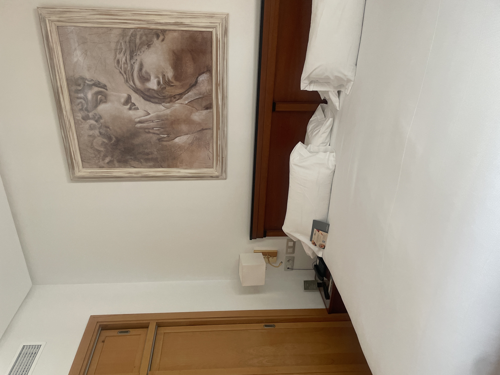 The image depicts a hotel room in Seville, Spain. The room features a large, made bed with white linens and pillows. Above the headboard hangs a framed painting of two classical figures. To the left of the bed, a nightstand holds a telephone and a lamp. Adjacent to the nightstand is a tall, wooden wardrobe. The walls are painted white, and a ventilation grate is visible near the ceiling. The overall impression is one of a well-appointed and comfortable hotel room.