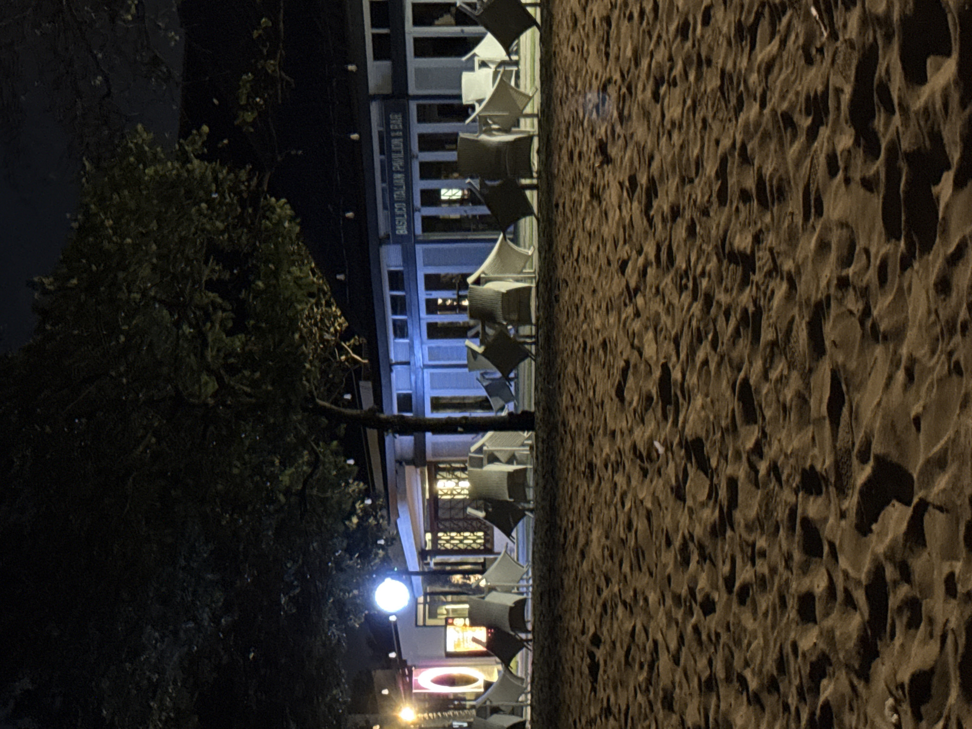 The image depicts a nighttime scene on a sandy beach in Denpasar, Indonesia. In the background, a building with the sign "BASILICO ITALIAN PAVILION & BAR" is illuminated, suggesting it is a restaurant or bar. Several white chairs are arranged on the sand in front of the building, some appearing to be outdoor seating for the establishment. The beach is covered in sand with footprints and textured surfaces. Trees with dark foliage are visible in the foreground and middle ground, casting shadows. Lighting fixtures are also present, including a bright circular light near the building and string lights further back. The overall atmosphere is dark and quiet, indicating the late hour.