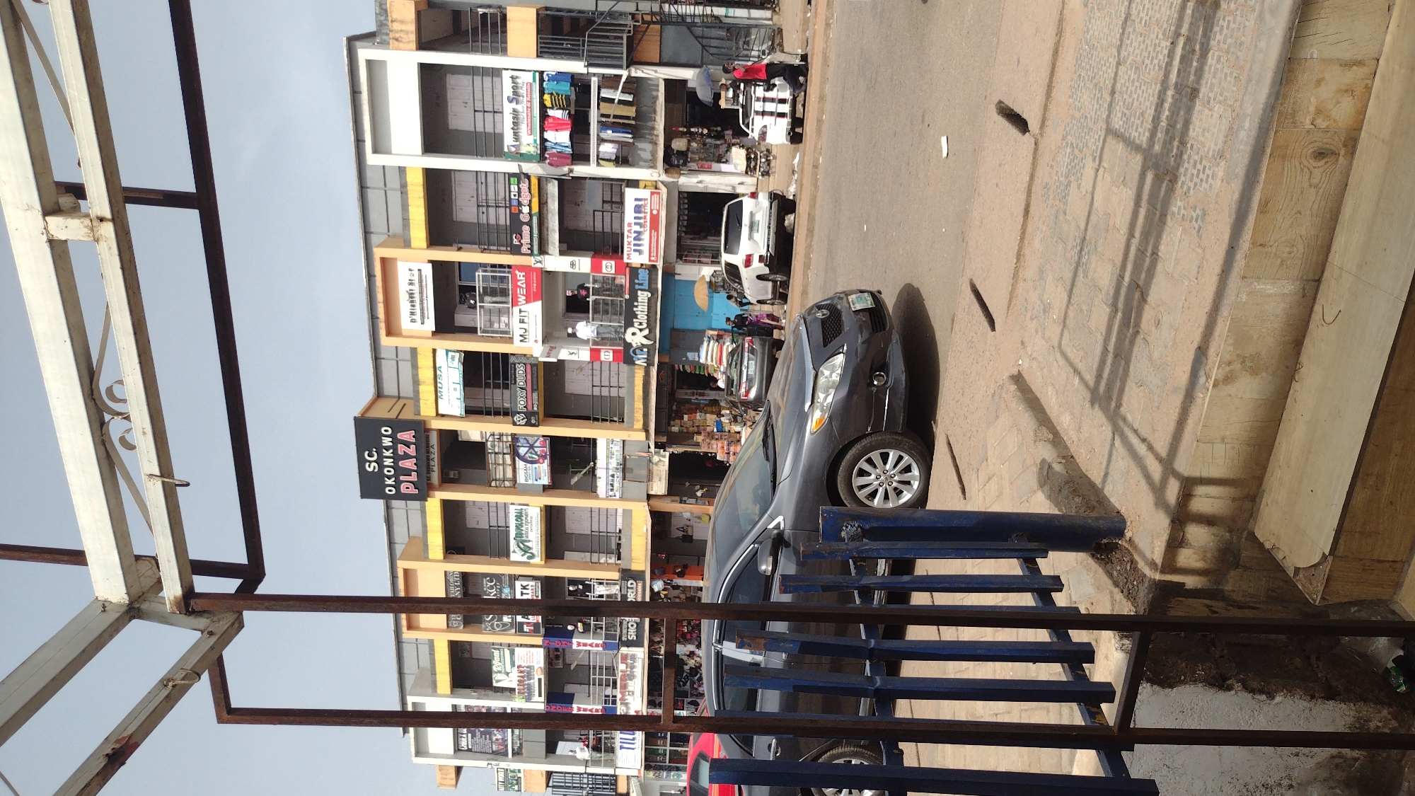 The image shows a street scene in Jos, Nigeria, in front of the "SC. Okonkwo Plaza", a multi-story building with various businesses. There are signs for "Muntasir Sport," "Prime Gadgets," "Foxy Duds," "MJ FIT WEAR," "Muktar Jinjiri," and "KP Clothing Line" among others, suggesting a commercial area. Several cars are parked along the street, including a dark-colored sedan and a white SUV. People are visible walking on the sidewalk. A blue metal railing and a partial view of a metal structure frame the foreground. The street is paved and appears to be in a state of moderate use.