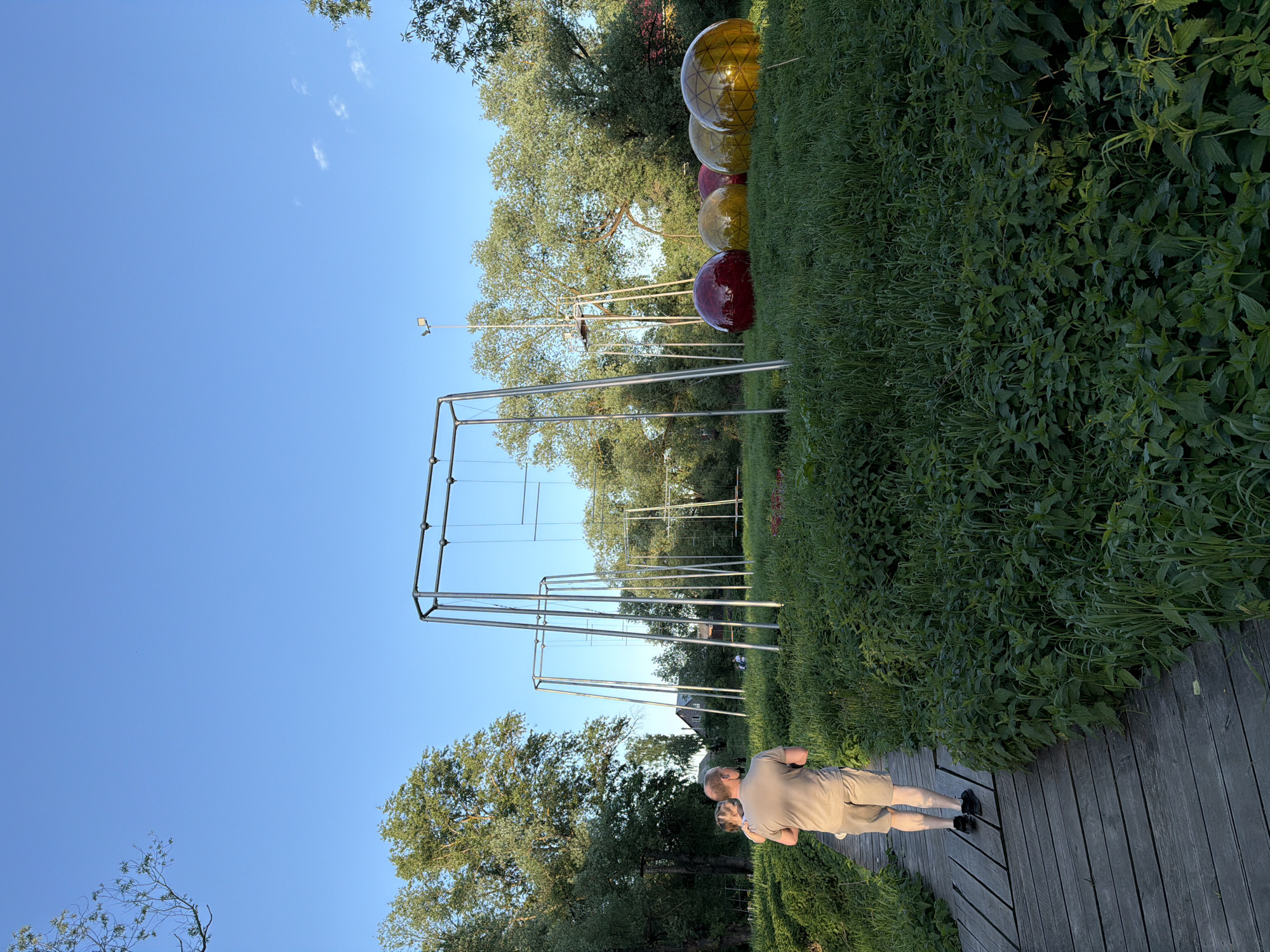 The image depicts an outdoor scene in Sin'kovo, Russia. In the foreground, a wooden boardwalk is visible, with a man standing on it, appearing to be taking a photograph. The man is wearing tan shorts and a shirt, and black shoes. Next to the boardwalk is a patch of dense green vegetation. Behind the vegetation is a metal structure with vertical and horizontal metal bars, resembling a frame or an incomplete building. Beyond this structure are several large, colorful, translucent spheres (red, yellow, and gold) that seem to be part of an art installation or garden decor. Trees with green foliage are scattered throughout the scene, under a clear blue sky. A street lamp is also visible.