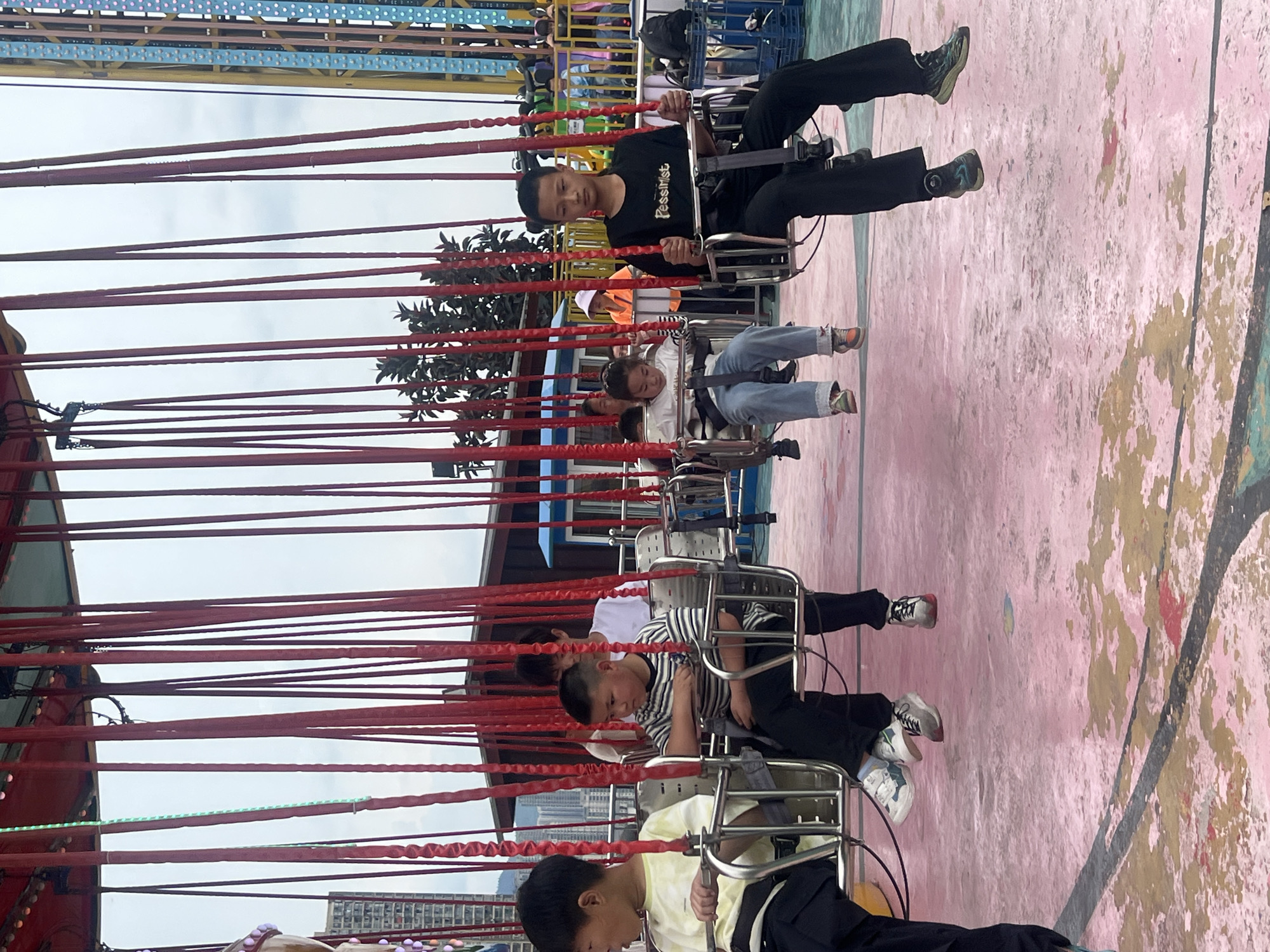 A group of people are riding a swing carousel ride. The ride has red chains suspending the seats. The background shows buildings, trees, and other parts of the amusement park. The flooring is painted in pink and brown. The location is in Nan'an District, China.