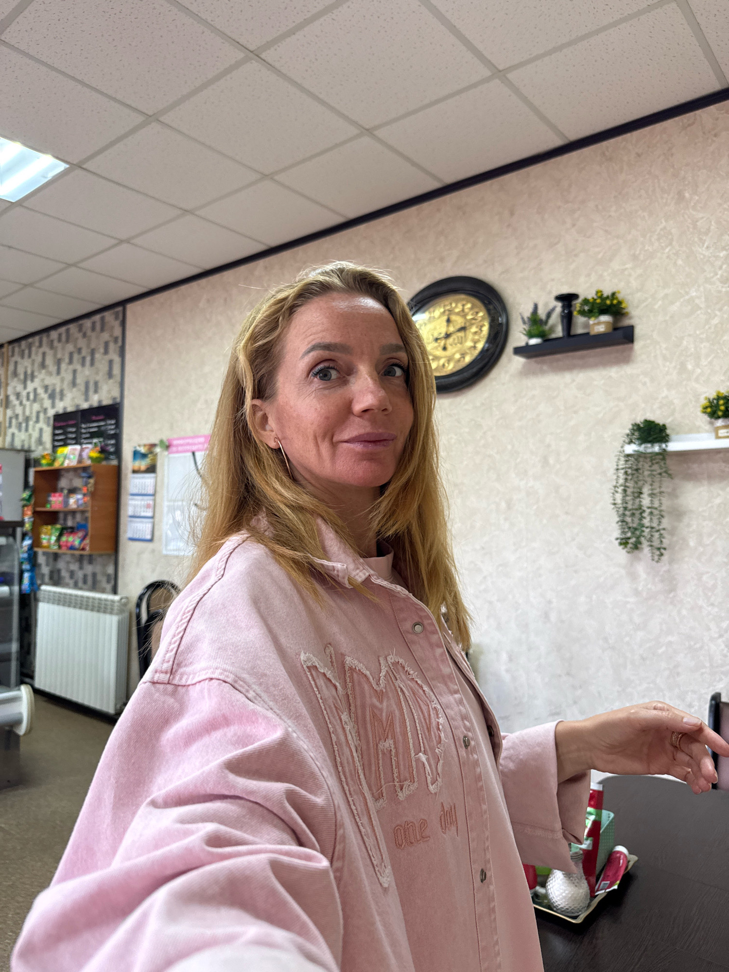A woman with blonde hair, wearing a pink denim jacket, is standing in what appears to be a small convenience store or cafe. The background shows shelves stocked with various items, a radiator, a clock on the wall, and decorative plants on shelves. The woman is looking towards the camera, and her right hand is resting on a dark surface. The scene is indoors with fluorescent lighting. The location is identified as Ryabovo, Russia.