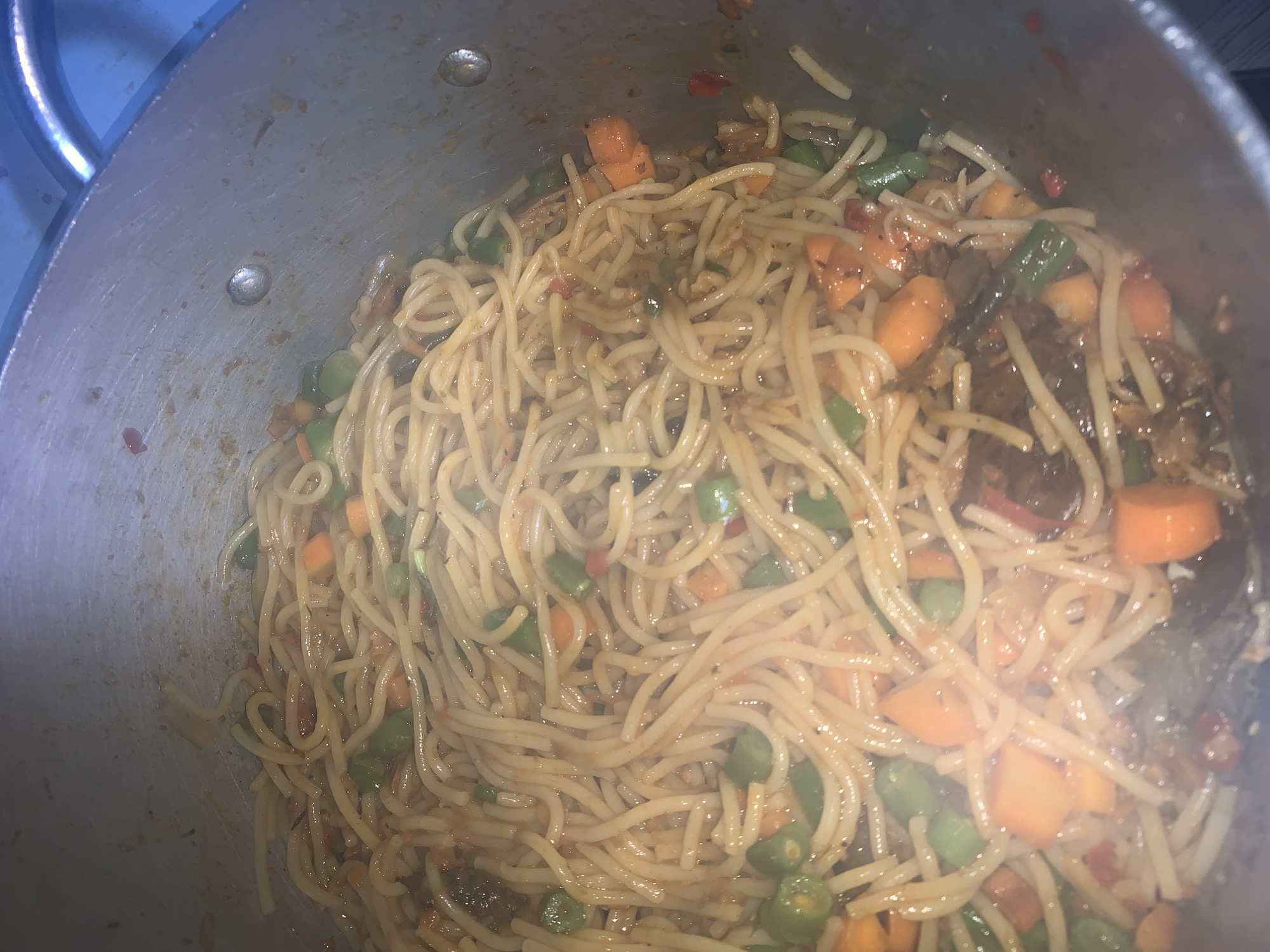 A close-up, overhead shot shows a pot filled with spaghetti, diced carrots, and green beans in a savory sauce. The spaghetti is thin and appears to be cooked al dente, with a glossy coating from the sauce. The carrots are bright orange and cut into small cubes, interspersed with the vibrant green of the green beans. The sauce has a reddish-brown hue and seems to contain some finely chopped ingredients. The pot itself is metallic and appears to be made of aluminum or stainless steel, with some visible smudges and cooking residue on its inner surface. The lighting is somewhat dim, but highlights the textures and colors of the food. The image is taken from a high angle, looking directly down into the pot. There is no visible indication of location in a city, nor any people or actions.
