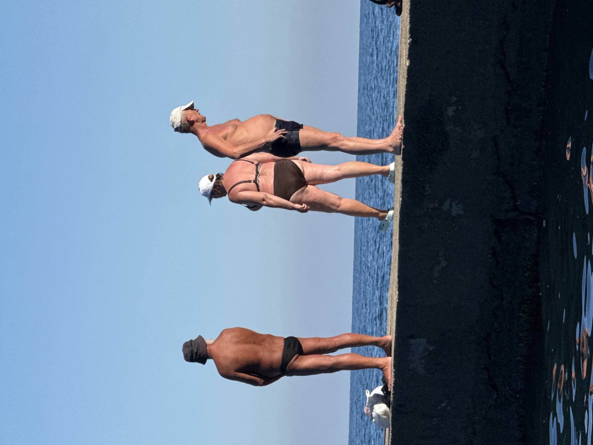 Three individuals are standing on a concrete pier with the sea and clear blue sky in the background. The person on the left is a man wearing a dark bucket hat and black swim briefs. He is facing away from the camera. The person in the middle is a woman wearing a white cap, a bikini with a patterned top and dark bottoms. She is facing to the right. The person on the right is a man wearing a white baseball cap and dark swim trunks. He is facing to the right, with his left hand on his hip. Two white bags are on the pier near the person on the left. The scene appears to be in Partenit, Ukraine.
