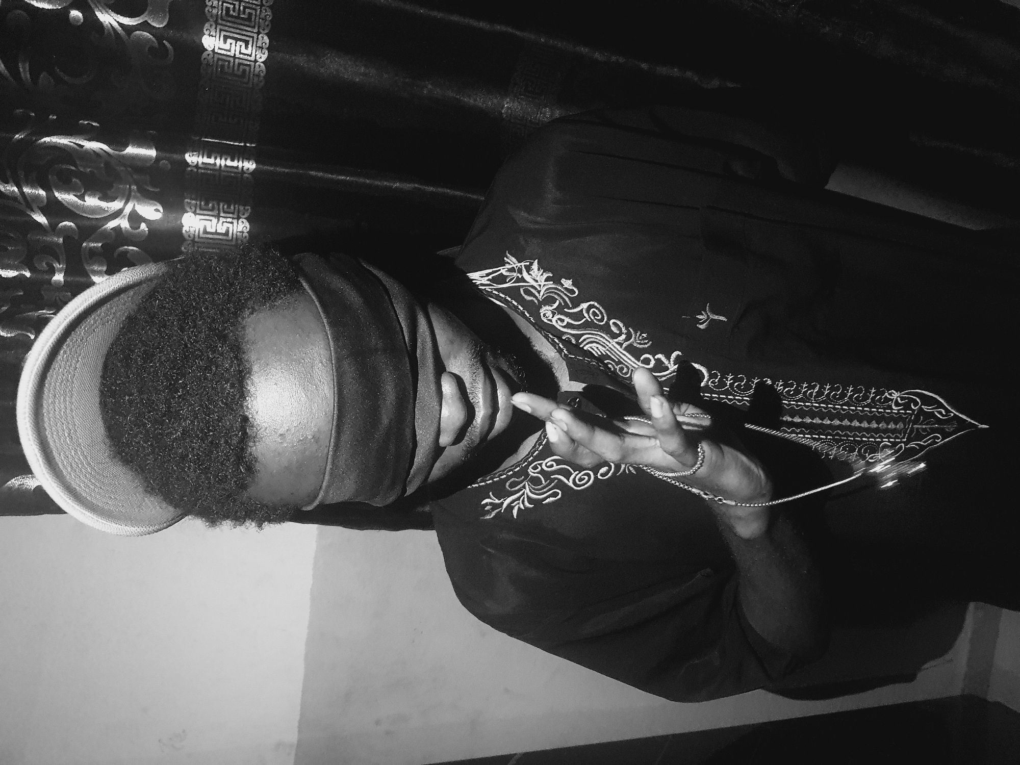 A person with dark skin and hair is wearing a black shirt with ornate white embroidery around the neckline and chest, and a light-colored hat. A dark cloth covers their eyes. They are holding a delicate chain in their hands. The location is unknown but presumed to be in the city of Lokoja, Nigeria. The background includes a light-colored wall and a dark curtain with a metallic pattern.