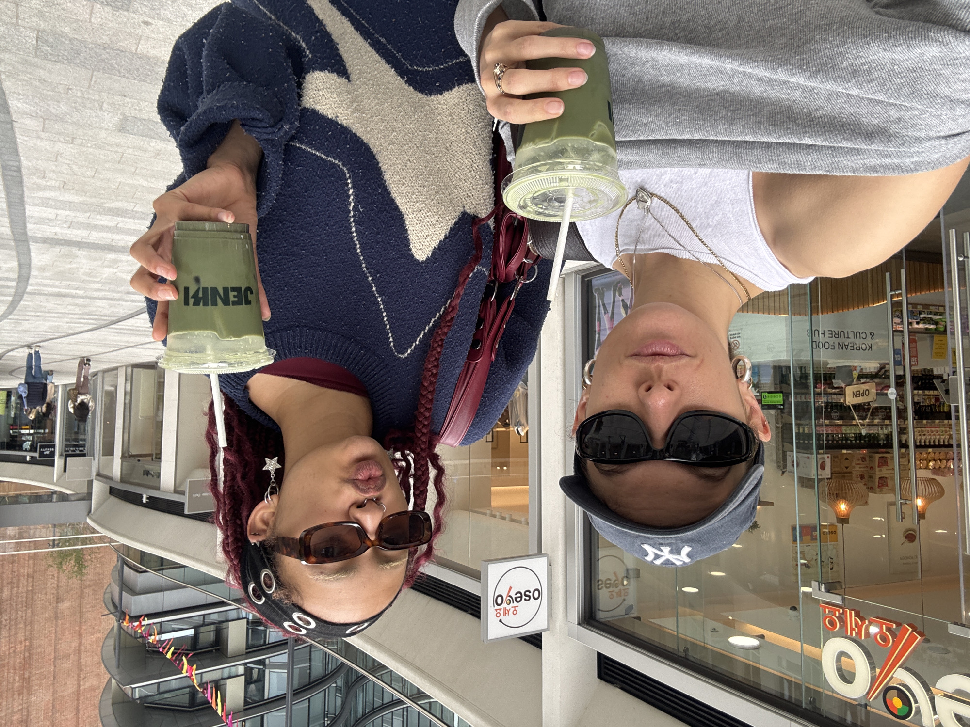 Two individuals are posing for a photo while holding green drinks outside a building in London, United Kingdom. The person on the left wears a navy sweater, red top, dark sunglasses, and a black headband. Her hair is red, and she is holding a green drink with the name "JENIFI" printed on the cup. The person on the right wears a gray top, white shirt, and dark sunglasses, along with a gray baseball cap. She is also holding a green drink with a straw. A shop named "OSEVO" is visible in the background, along with text indicating "KOREAN FOOD & CULTURE HUB" and an "OPEN" sign. The surrounding environment includes parts of modern buildings, street decorations, and reflections of the street scene in the shop windows.