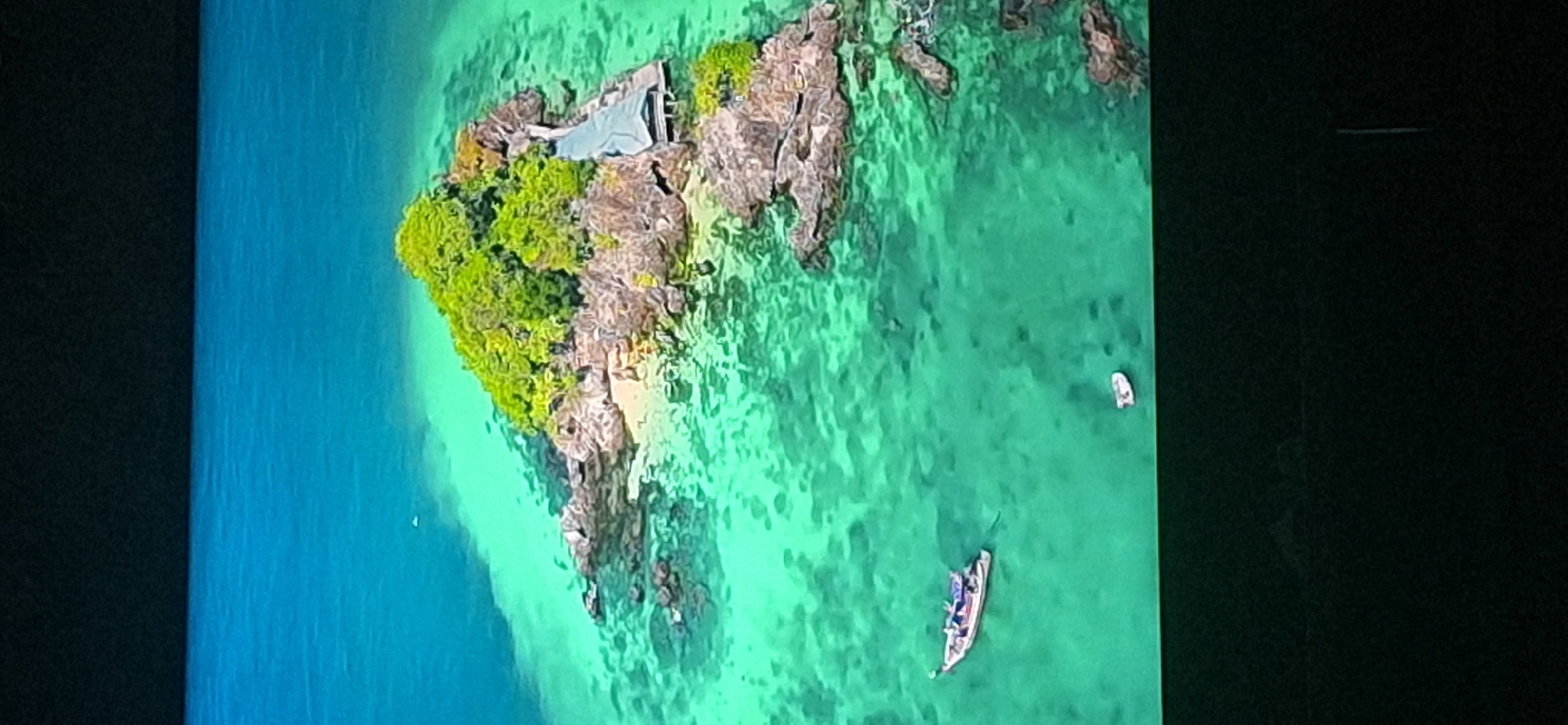 An aerial view shows a small, rocky island with lush green vegetation in the center. A building with a light-colored roof and wooden walkway is built on the right side of the island. The island is surrounded by clear, turquoise water. Two boats are visible in the water near the island: a larger boat with several people on board is to the left, and a smaller boat is further to the right. The bottom of the image is black, obscuring any view of the surrounding land or city. There is no indication of the location being Bila Tserkva, Ukraine.