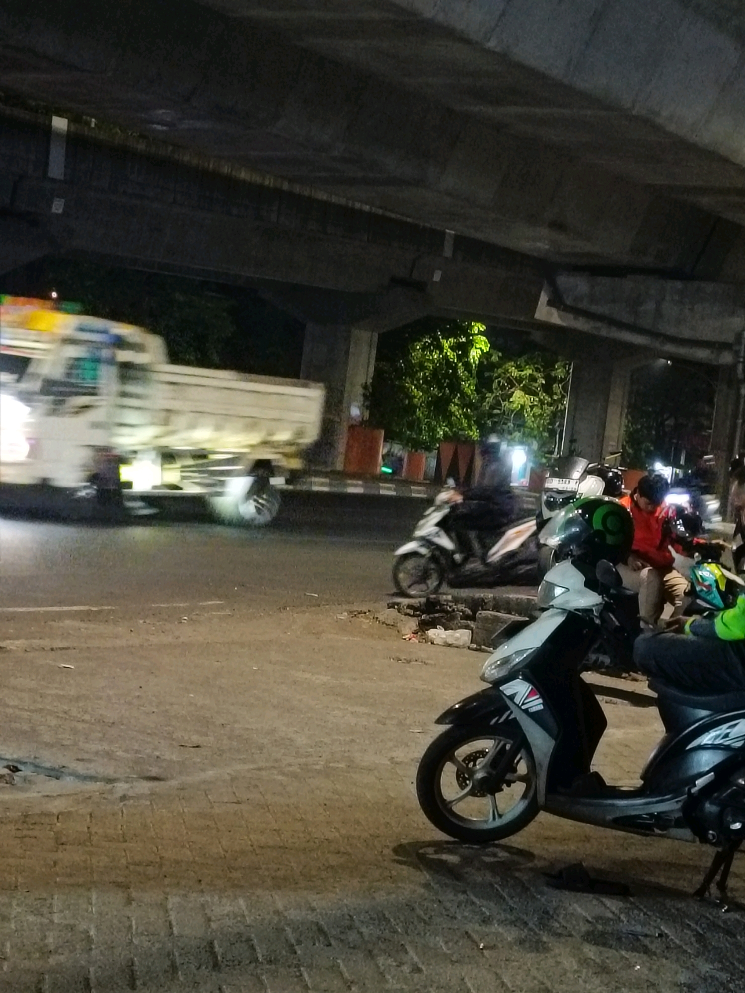 The image depicts a dimly lit scene at night under an elevated structure, likely a flyover or bridge, in Makassar, Indonesia. In the foreground, a parked black and white scooter is visible with its front wheel turned. Further back, several people are gathered around other parked motorcycles. One person in a red jacket is seated, looking down at their phone, while others stand or sit nearby.
To the left, a white truck with a flatbed is in motion, captured with motion blur. The truck has bright headlights and appears to be driving on a paved road. The environment includes elements of urban infrastructure such as concrete pillars supporting the elevated structure, planters with greenery, and a sidewalk made of interlocking bricks. The overall atmosphere is one of a street-level scene at night, possibly a gathering spot or a waiting area for transportation.