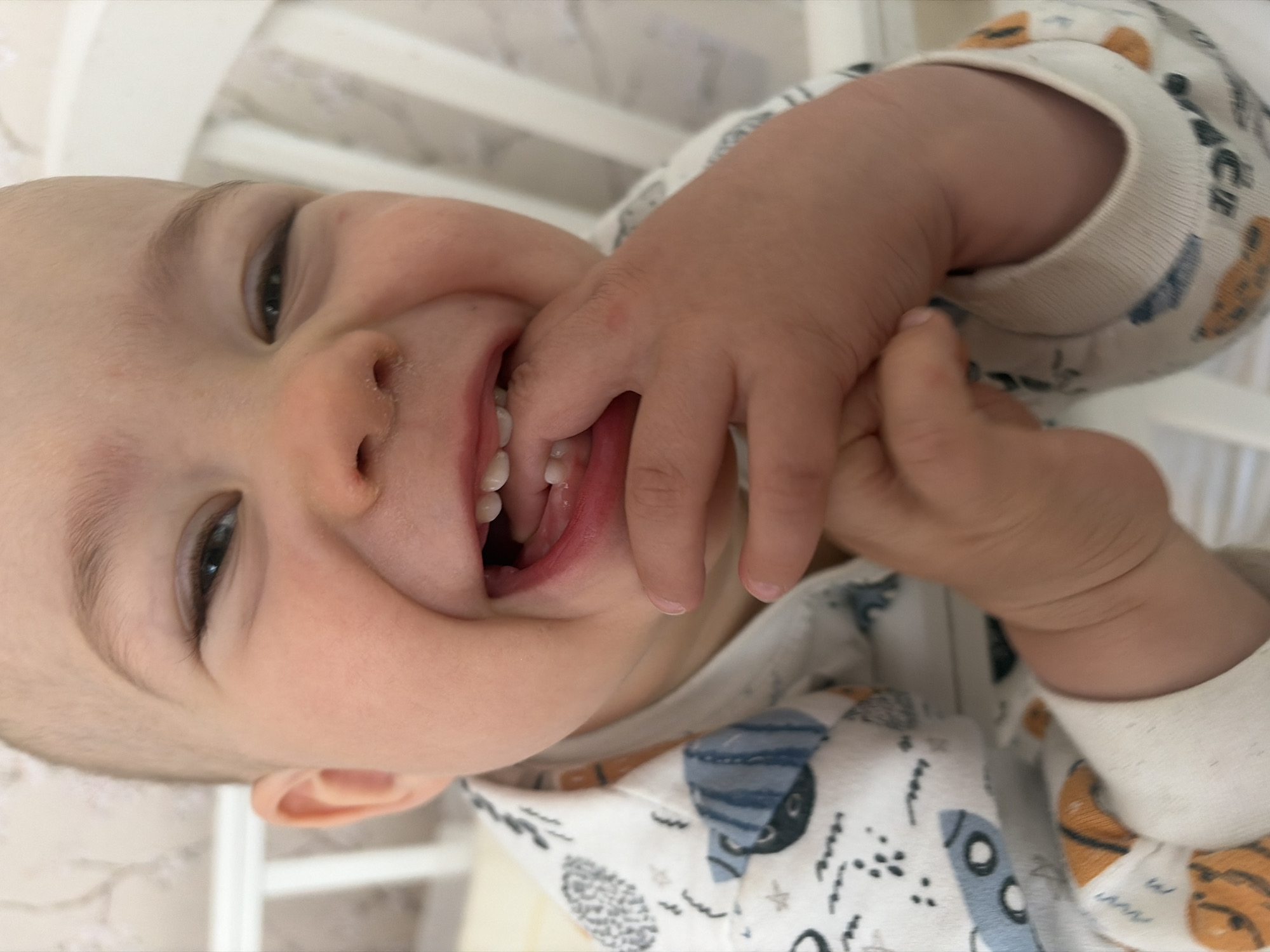 A close-up shot of a baby lying on their back, positioned in a white crib. The baby is wearing a white onesie with a print of vehicles and animals. Their face is the primary focus, with a bright smile revealing their upper and lower teeth. The baby's hand is raised, with their fingers partially in their mouth. The background consists of the vertical slats of the crib and a blurred surface behind the crib, suggesting a room environment. There is no location information regarding Chaikovsky, Russia.
