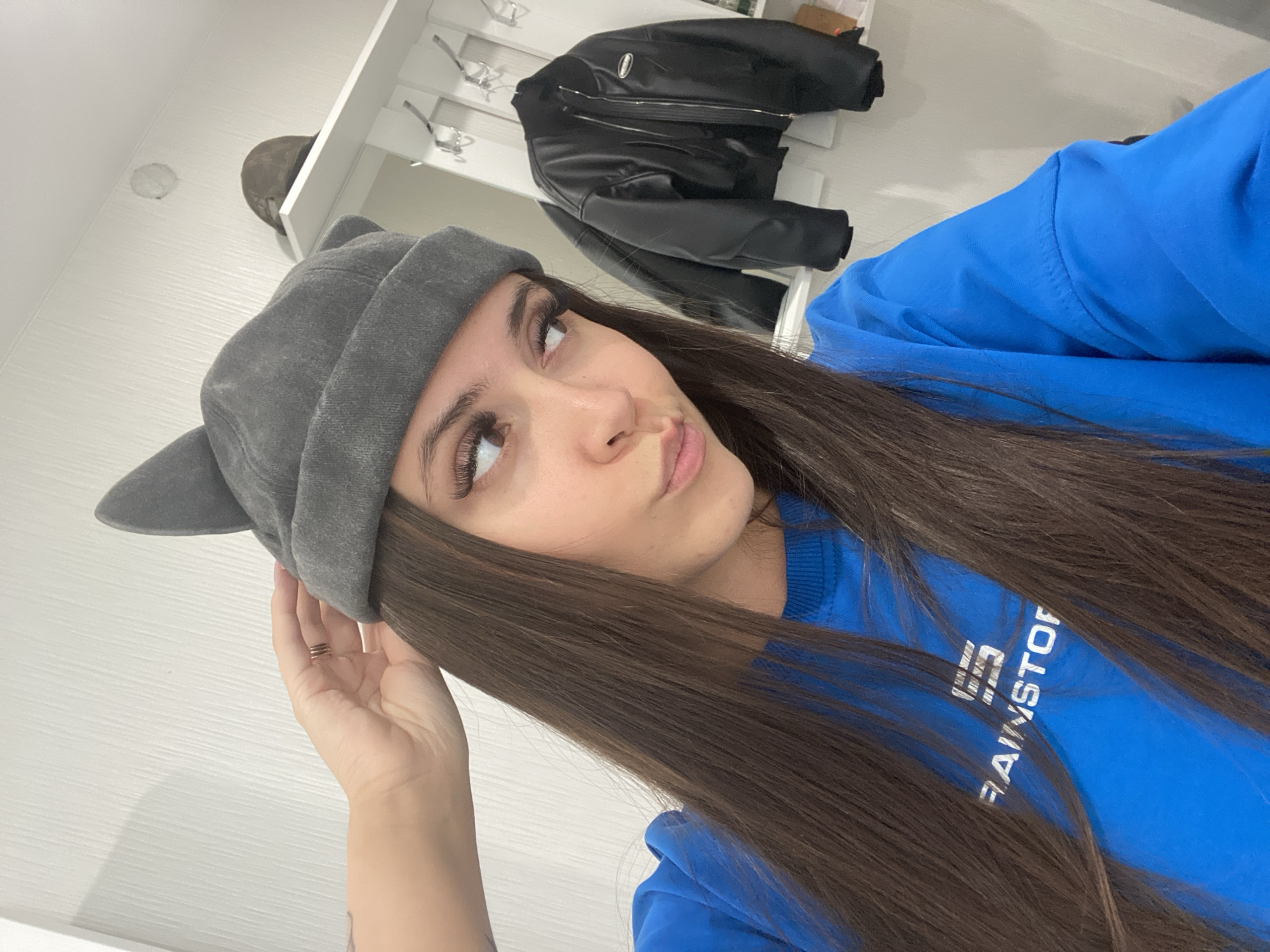 The image shows a young woman with long, dark hair, wearing a grey beanie with cat ears and a bright blue long-sleeved shirt. The shirt has white text on the sleeve that reads "RAINSTOP". She is looking up and to the side with a slightly pouting expression, her hand is resting on her head near the beanie. The background appears to be an interior setting, possibly a hallway or closet, with a white paneled wall and a white shelf unit. On the shelf unit, a black leather jacket is hanging. There is also a glimpse of a brown hat on a shelf. The lighting suggests it is daytime indoors. The woman's makeup is visible, including dramatic eyelashes.