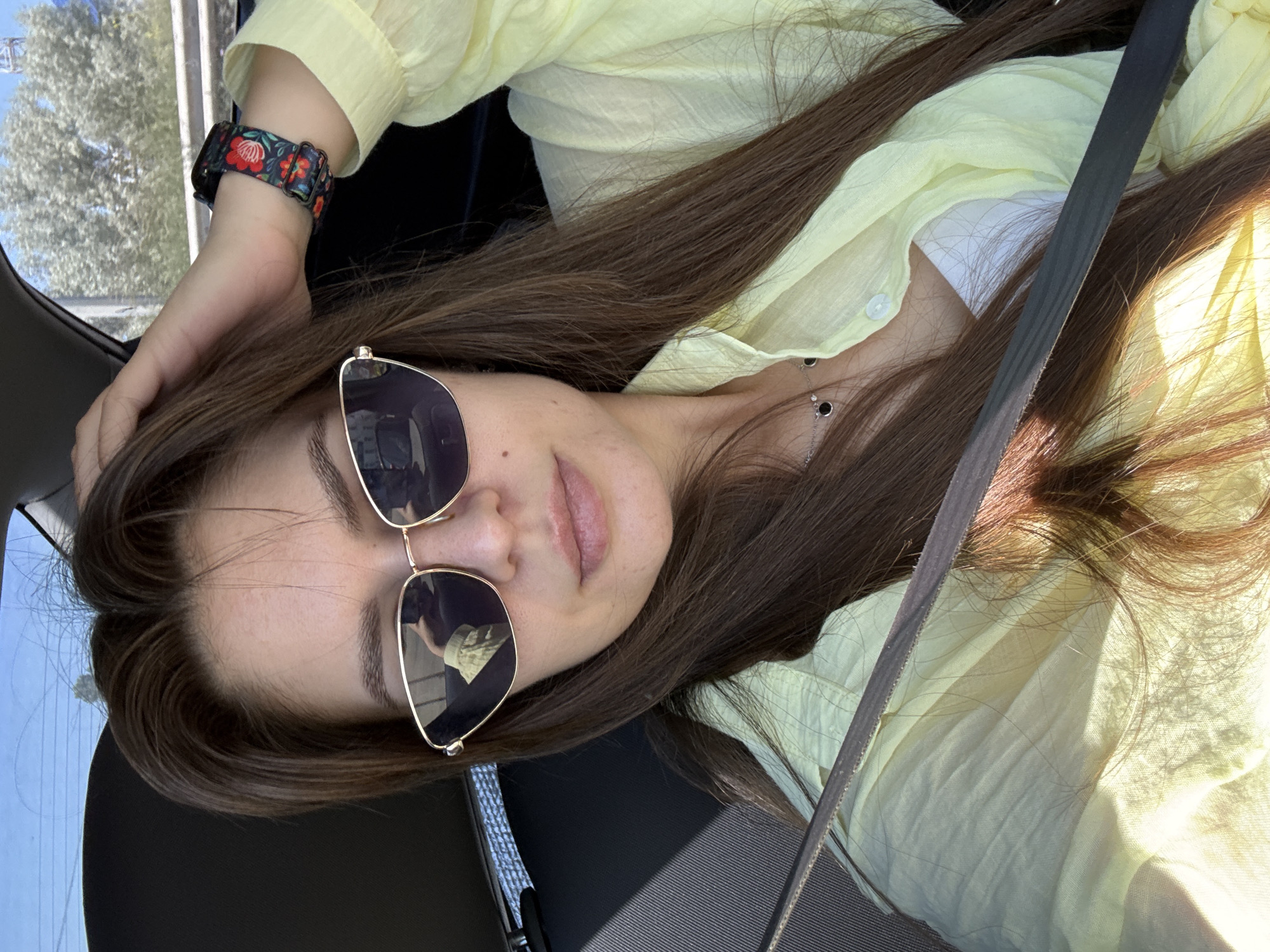 A close-up selfie shows a woman with long brown hair wearing sunglasses and a light yellow shirt. She is in a car, and the seatbelt is visible across her chest. Her right hand is resting on her head, and she is wearing a floral-patterned watch on her left wrist. The background suggests an outdoor environment, possibly a city street or highway, indicated by trees and a clear sky visible through the car window.