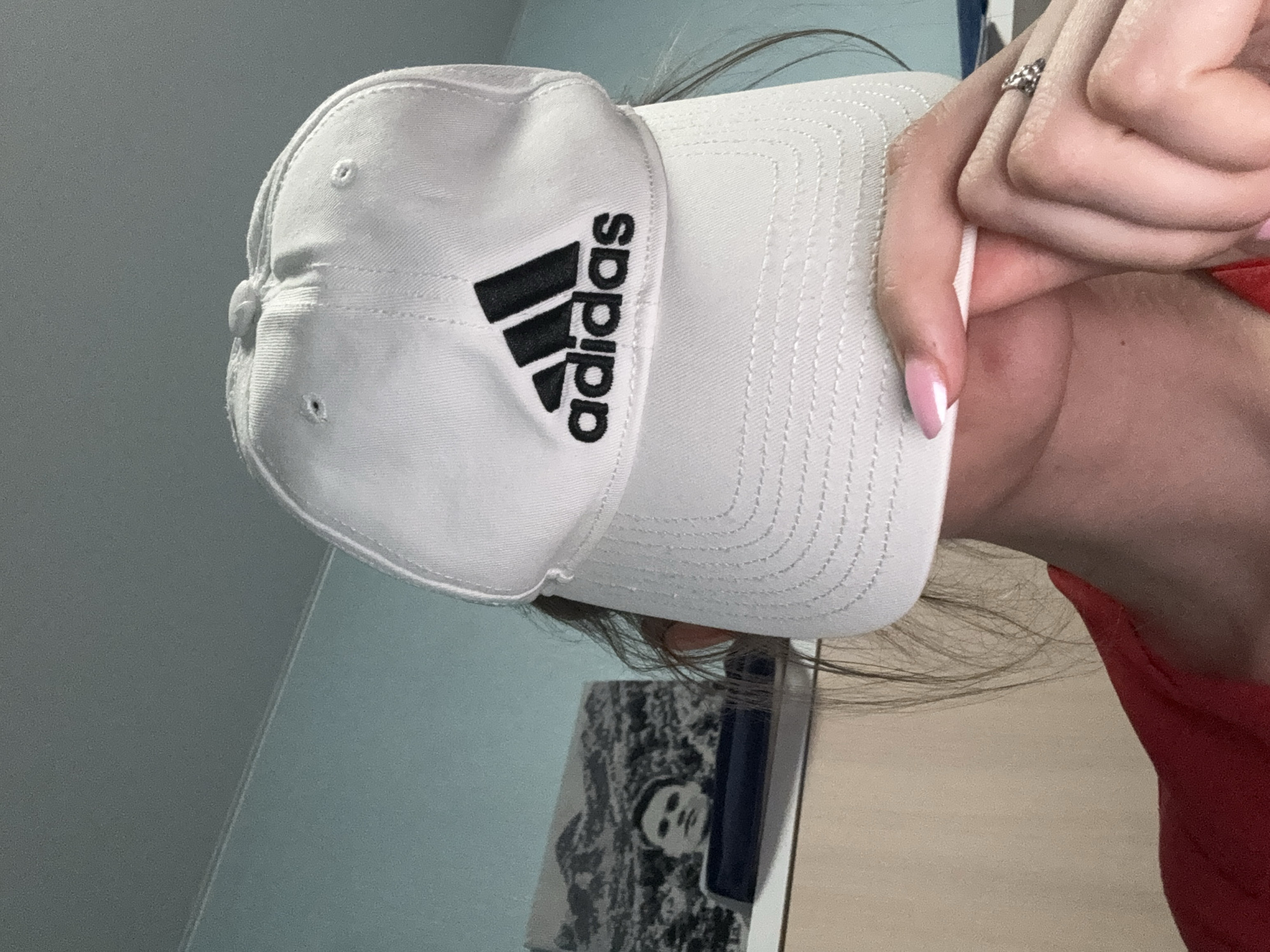 A person is wearing a white baseball cap with the Adidas logo. The person is wearing a ring on their finger and has pink fingernails. The background is a light blue wall and a wooden surface.