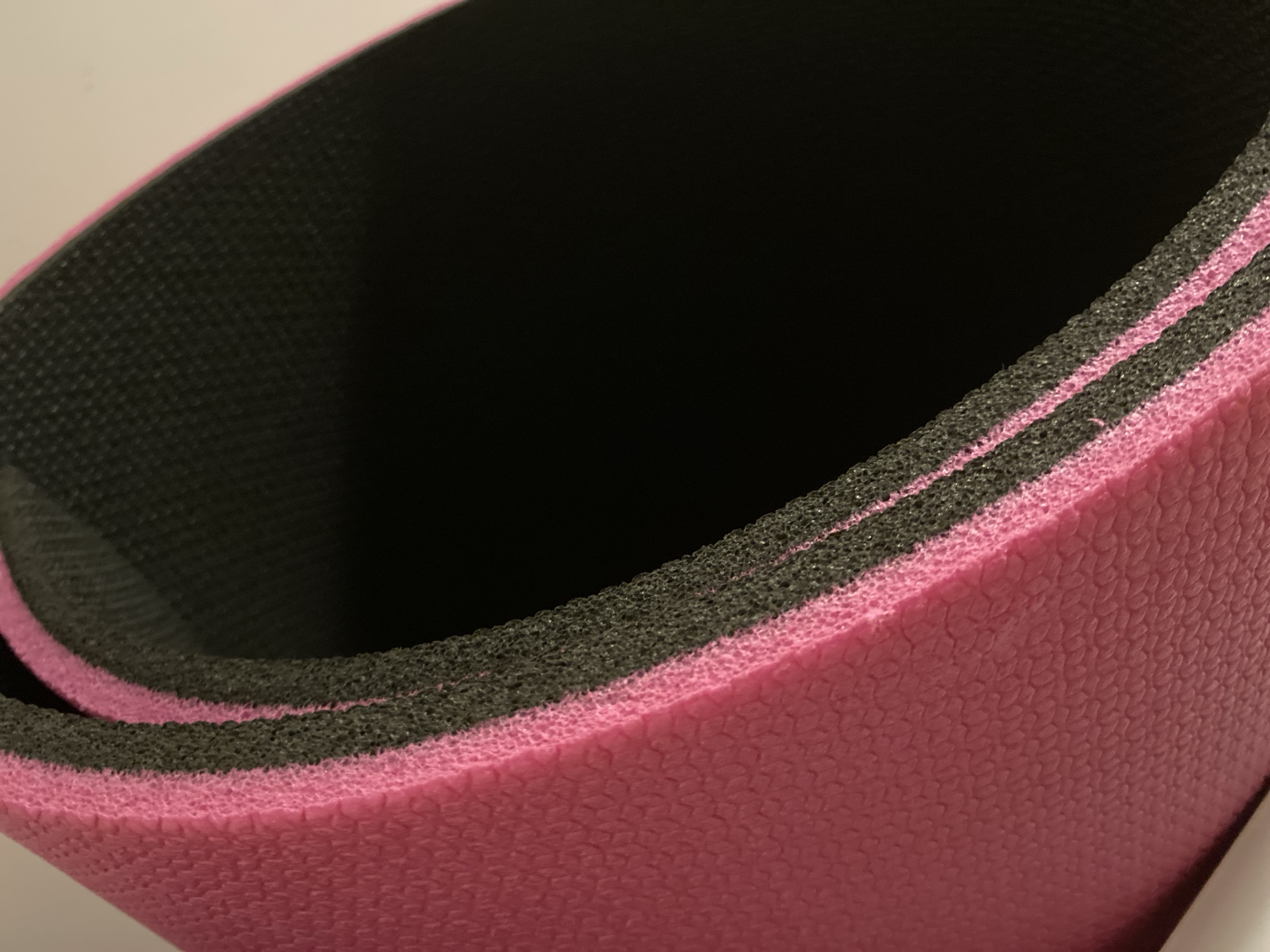 A close-up view of a partially rolled-up, two-layered yoga mat. The visible layers are pink and black. The pink layer has a textured surface. The black layer appears to have a porous texture. The item is situated on a light-colored surface. No people, actions, or specific environmental context (such as Alanya, Turkey) are identifiable from the image.
