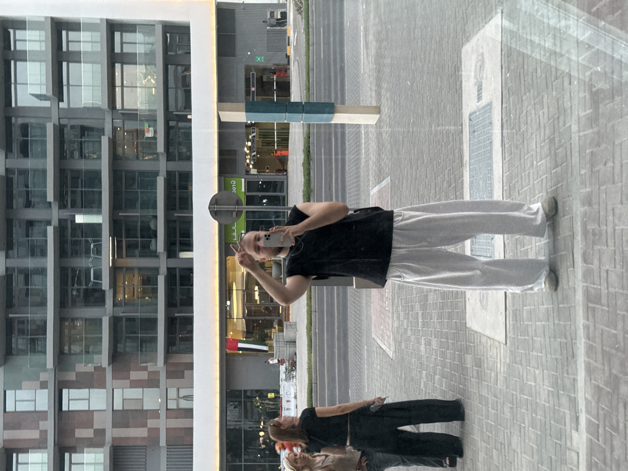 A person is taking a selfie with a phone while standing on a paved sidewalk in front of a modern building. The building has a facade of glass windows and textured panels. The person is wearing a black t-shirt and light gray pants, and they are making a peace sign with their hand. In the reflection on the building's glass, another person in a dark outfit is visible, along with a blurry reflection of what appears to be a street and a few trees. There are also reflections of lights and signs. The scene suggests an urban environment. The time of day appears to be daytime, and the weather seems clear. There is some Arabic text visible on a green sign in the reflection, and the UAE flag is also visible reflected.