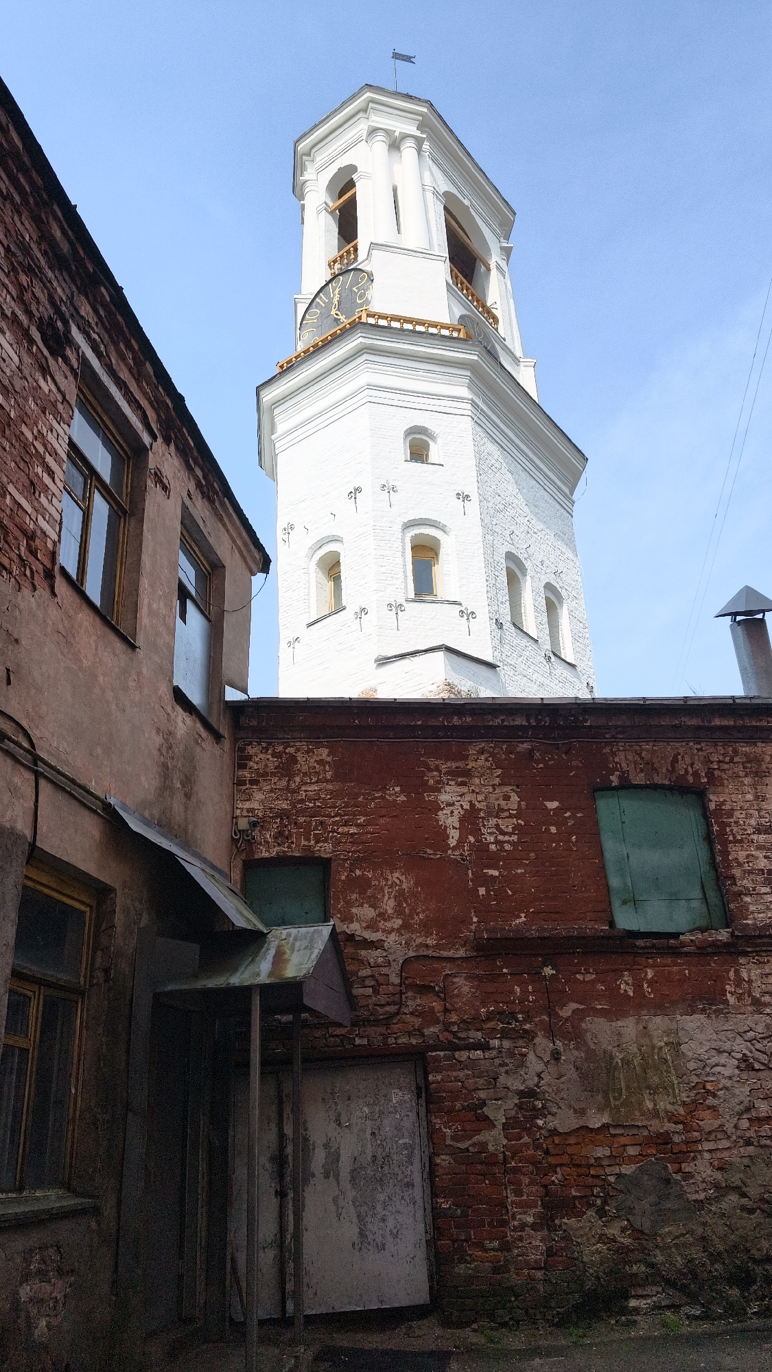 The image captures a low-angle view of a bell tower and surrounding buildings, likely in Vyborg, Russia. The dominant structure is a tall, white, octagonal bell tower with arched openings and a clock face adorned with gold Roman numerals. The tower features intricate decorative elements above the clock and smaller windows with yellow-tinted glass. Below the tower, red brick buildings are visible. One building on the left has a weathered, reddish-brown facade with several windows, some of which appear to have reflective surfaces. Another building in the foreground, also constructed of red brick, shows signs of decay and peeling paint. This structure has a dark, weathered door beneath a metal awning. A green shuttered window is also present on this building. The sky is a clear, pale blue. The scene conveys a sense of age and perhaps neglect, with the stark white of the tower contrasting with the rough textures of the brick structures.