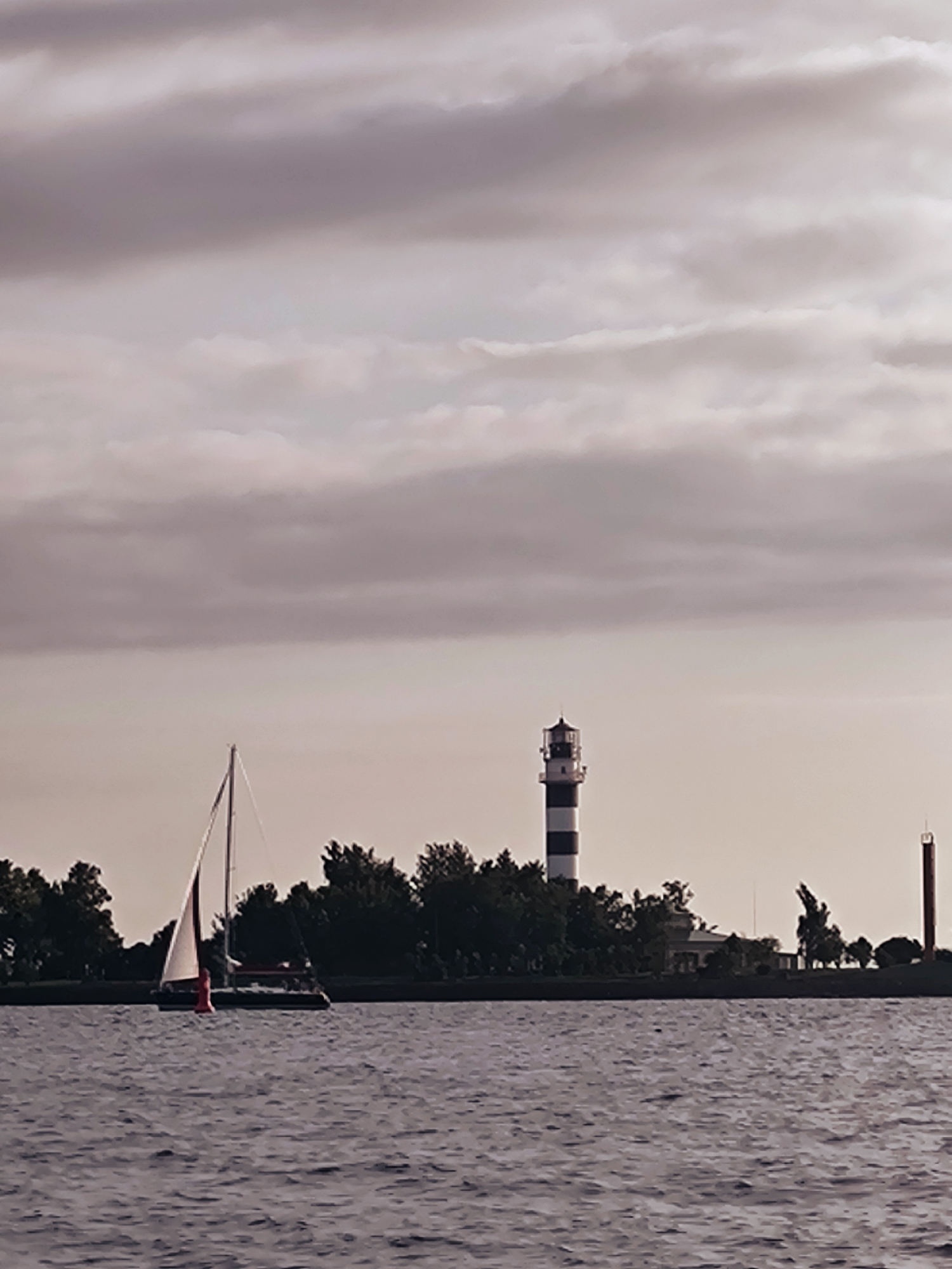 The image depicts a serene waterscape with a lighthouse and a sailboat in the distance. The sky is overcast with soft, muted colors. On the right side of the image, a tall, striped lighthouse stands prominently, its black and white bands clearly visible. To the left of the lighthouse, a sailboat with a white sail is gliding across the water. Trees line the shore behind the lighthouse and sailboat, forming a dark green silhouette against the pale sky. The water is calm, with gentle ripples reflecting the diffused light. The overall atmosphere is tranquil and slightly melancholic, evoking a sense of quietude and natural beauty. The scene is likely located in Riga, Latvia, given the characteristic coastal landscape and presence of lighthouses that dot the Baltic Sea.