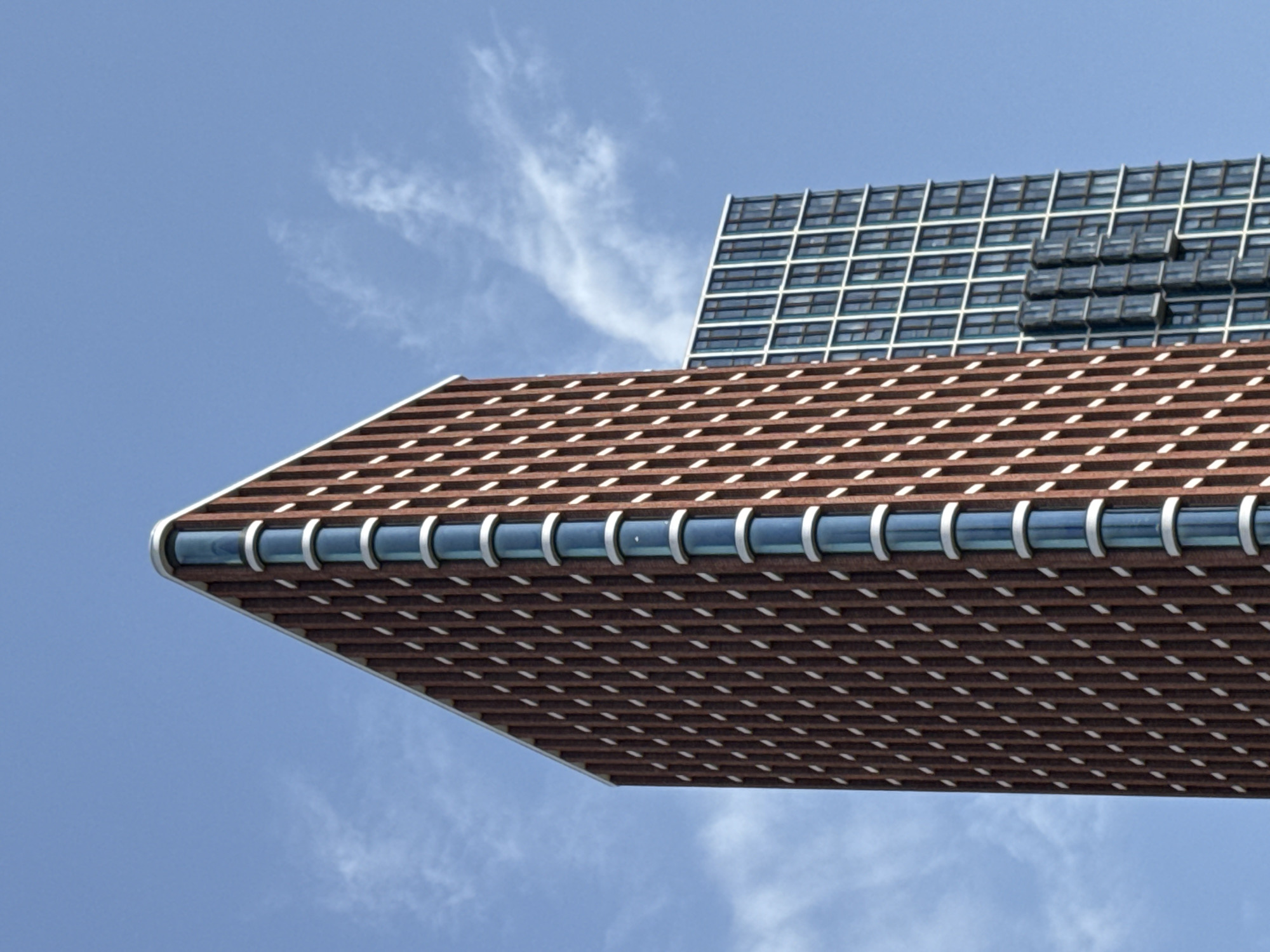 This is a low-angle, medium shot of two modern skyscrapers against a bright blue sky with scattered white clouds. The skyscraper in the foreground, on the left, is tall and angular, with a reddish-brown brick facade. It features a series of vertical white elements and narrow, curved, blue-tinted windows. The other skyscraper, further back and on the right, has a more regular, grid-like facade with numerous square, blue-tinted windows. The image captures the architectural details of both buildings and the contrast in their design. There is no indication of people or activity. The scene appears to be a contemporary urban environment.