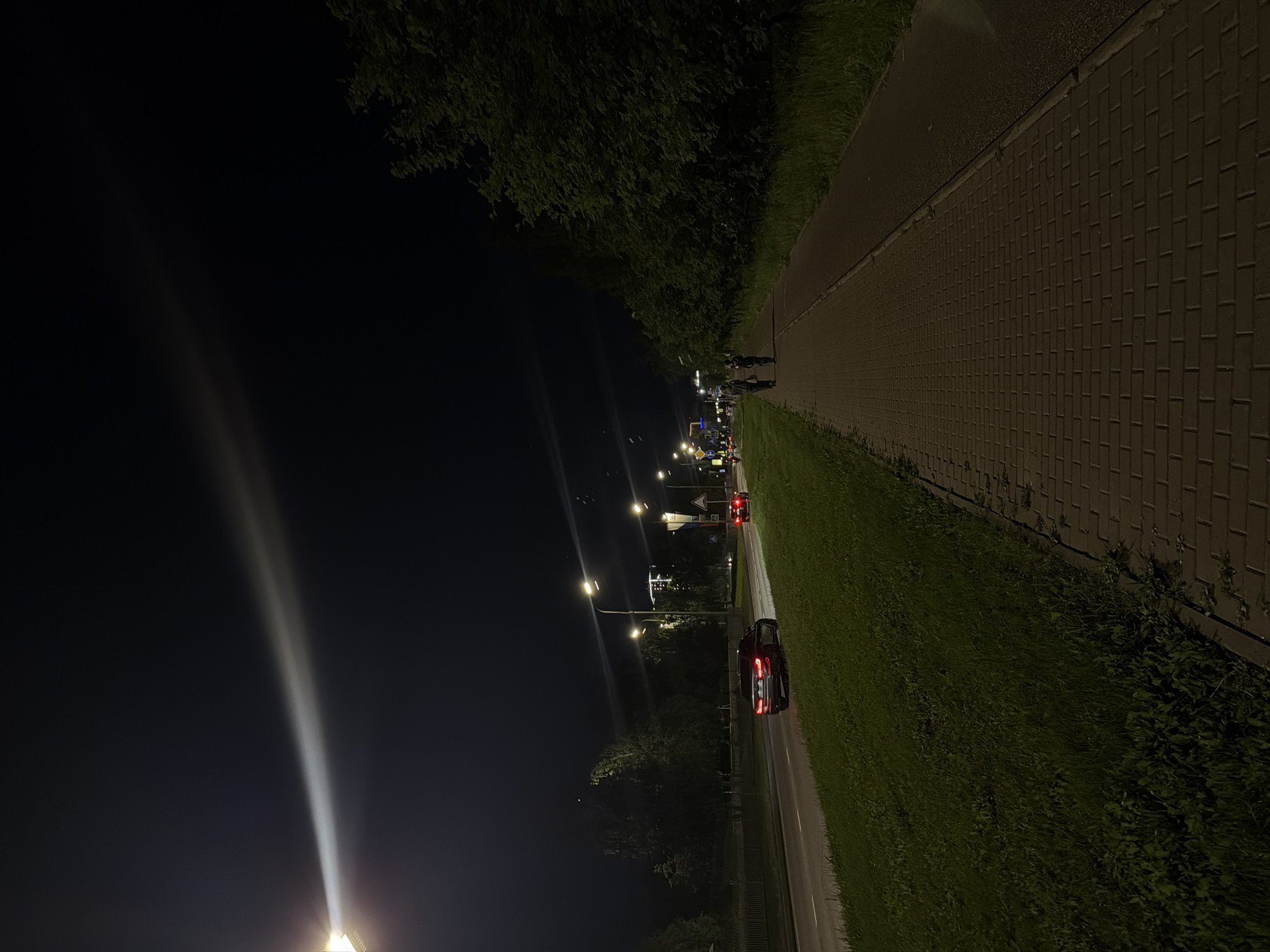 An image captures a nighttime scene on a street in Naberezhnye Chelny, Russia. A car is seen driving on a road to the left, illuminated by streetlights. Across the road, a paved pedestrian walkway runs alongside a grassy verge. Two individuals are walking on the walkway, heading away from the camera. Trees line the right side of the walkway, forming a dark, dense border against the night sky. A prominent streetlight casts a bright beam of light upwards and across the dark expanse of the sky.