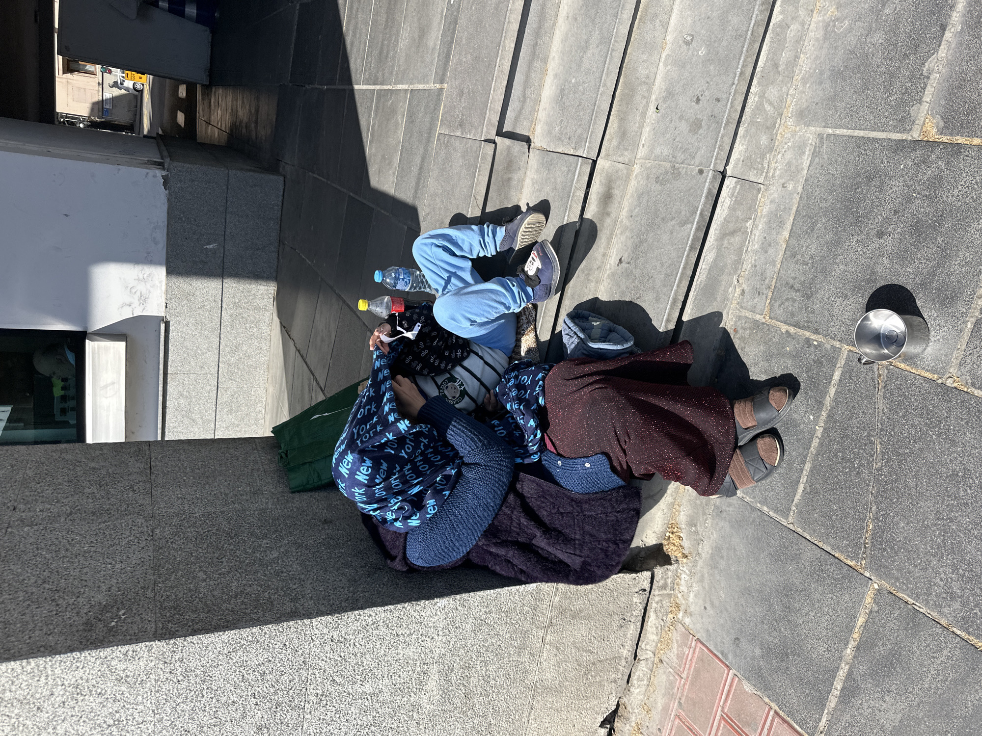 The scene is an outdoor shot from an elevated perspective, showing a group of people lying on a sidewalk with a metal collection cup nearby. The sidewalk is paved with large gray tiles. The group consists of three individuals; one is wearing blue sweatpants and gray sneakers, lying perpendicular to the second person, who is draped in dark fabric and wearing sandals. The third person is lying on their side covered in a blue printed blanket or fabric and a dark blanket. Empty plastic bottles are visible near the person in blue. The background shows a street corner with building facades. The partial text "New York" can be seen on the blue blanket. The location is identified as Tbilisi, Georgia.