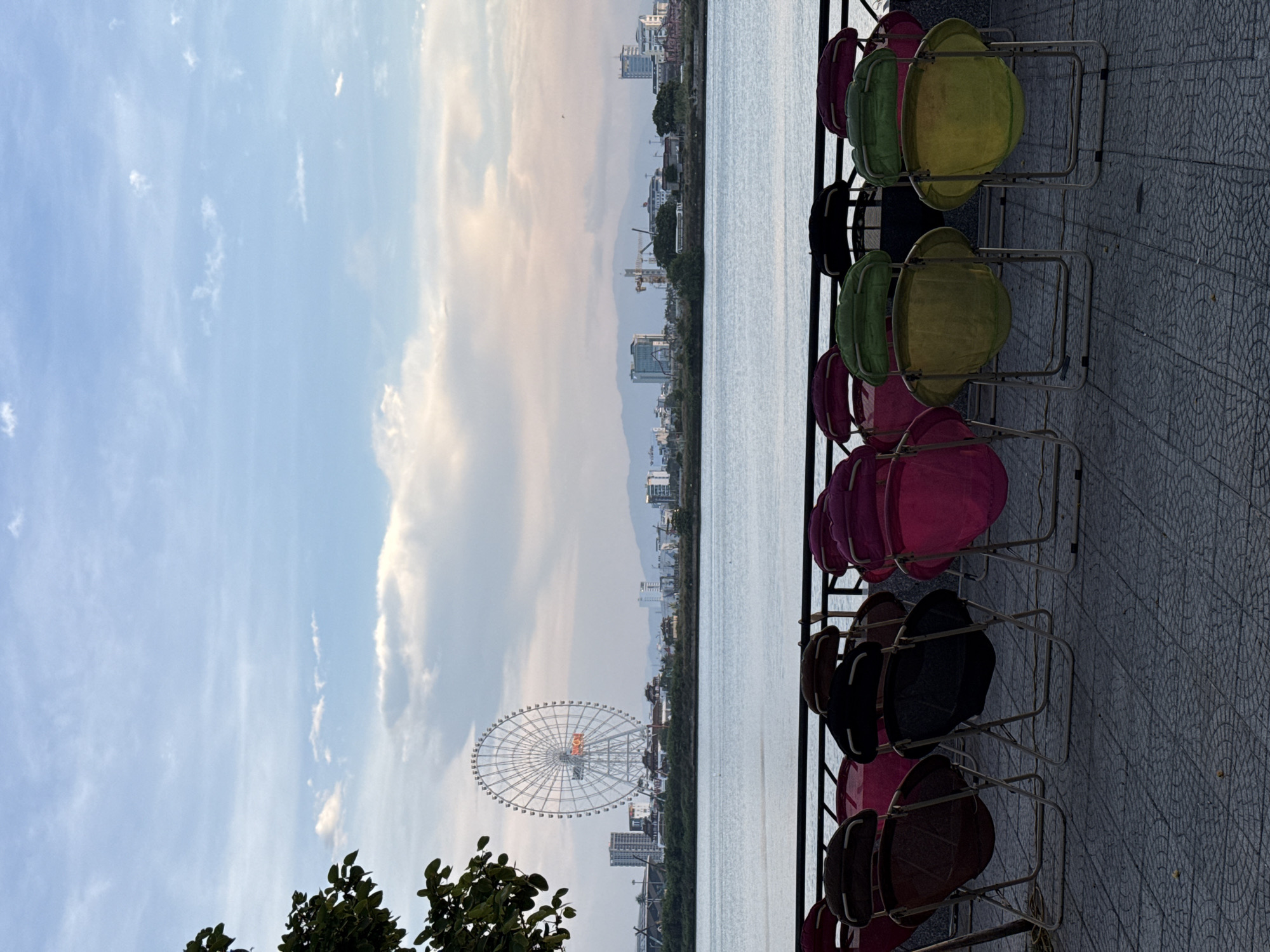 A scenic view of the Han River in Da Nang, Vietnam, is captured from a waterfront promenade. In the foreground, several colorful folding chairs are arranged, with some showing signs of wear. Beyond the chairs, the tranquil waters of the Han River stretch out, reflecting the sky. In the distance, the Da Nang city skyline is visible, dominated by the iconic Sun Wheel, a prominent Ferris wheel. The sky above is a soft blend of blue and pastel hues, with scattered clouds adding texture. The overall scene suggests a peaceful evening in the vibrant city of Da Nang.