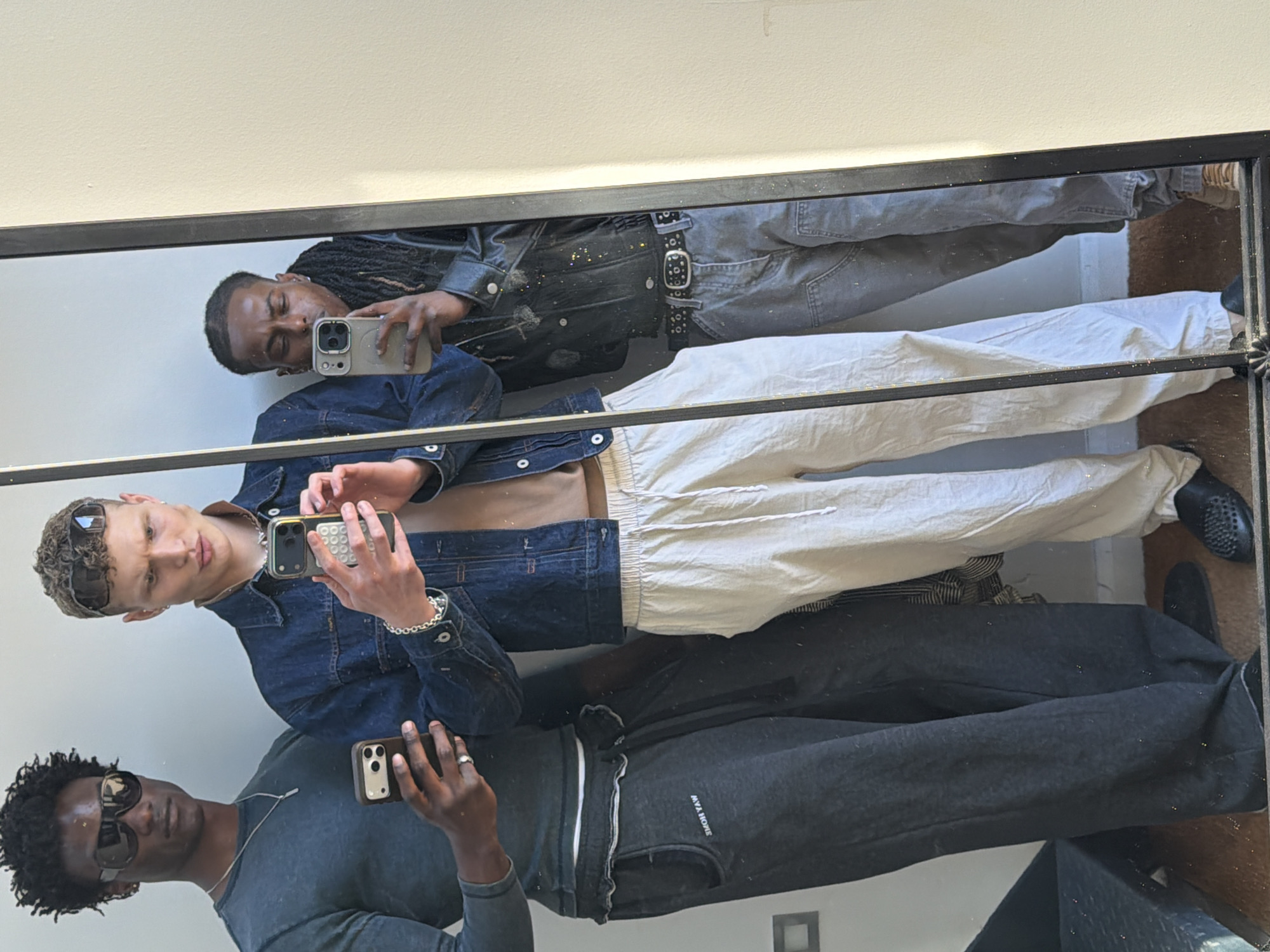 This image captures a mirror selfie featuring three men, standing upright as reflected in a large, dark-framed mirror. The indoor setting shows a light-colored wall reflected behind them.
On the viewer's upper left, a man with dreadlocks and a faded hairline is seen holding a light-colored smartphone. He is wearing a dark, possibly leather, jacket and dark pants, cinched with a prominent studded belt. His expression is neutral as he looks towards the camera.
In the center foreground, a man with short, curly hair wears dark sunglasses and a denim jacket, left open to reveal a bare midriff. He has several rings on his fingers and a silver bracelet on his wrist. He holds a smartphone with a distinctive white, textured case and a visible camera array. His pants are light-colored, possibly cream or off-white.
On the viewer's lower left, a third man with dark curly hair and dark sunglasses is visible. He is wearing a dark long-sleeved shirt and dark sweatpants. He holds a dark-colored smartphone and wears a silver chain necklace and a ring on his right hand. The mirrored text "MYA HOME" is visible on his sweatpants.
The overall activity is a group mirror selfie, with all three subjects looking towards the camera reflected in their phones. The lighting appears bright, suggesting daytime. Small, glitter-like particles are scattered across the mirror's surface, particularly visible on the dark frame.