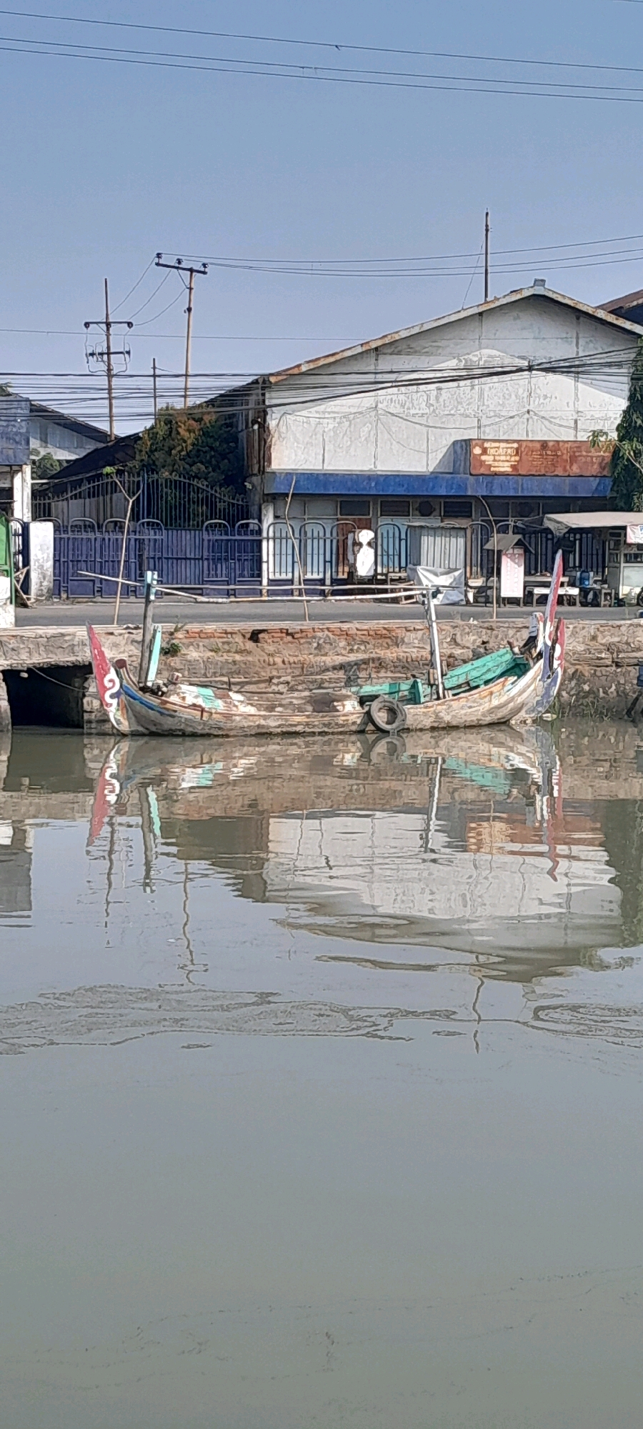 Two weathered, narrow boats are docked on a brick embankment beside a body of murky water. The boats are positioned parallel to each other, with the one on the left slightly closer to the foreground. Both vessels appear to be made of wood, with visible repairs and wear and tear. The boat on the left has a red and white flag at its bow. The boat on the right has a spare tire resting on its hull and a similar flag at its stern. The embankment is constructed of rough, reddish-brown bricks.
Behind the embankment, a street runs parallel to the water. On the far side of the street, a long, low-rise building complex stretches across the frame. The building has a weathered, light-colored facade with a blue awning and several arched window openings covered by white blinds. A metal fence, painted dark blue, separates the street from the building complex in some sections. Utility poles with electrical wires are visible against a clear blue sky. Power lines crisscross the upper portion of the image. The scene appears to be located in a commercial or industrial area, possibly near a harbor or canal in Pasuruan, Indonesia, given the style of the boats and buildings. The water reflects the buildings and boats, creating a mirrored image on its surface.
