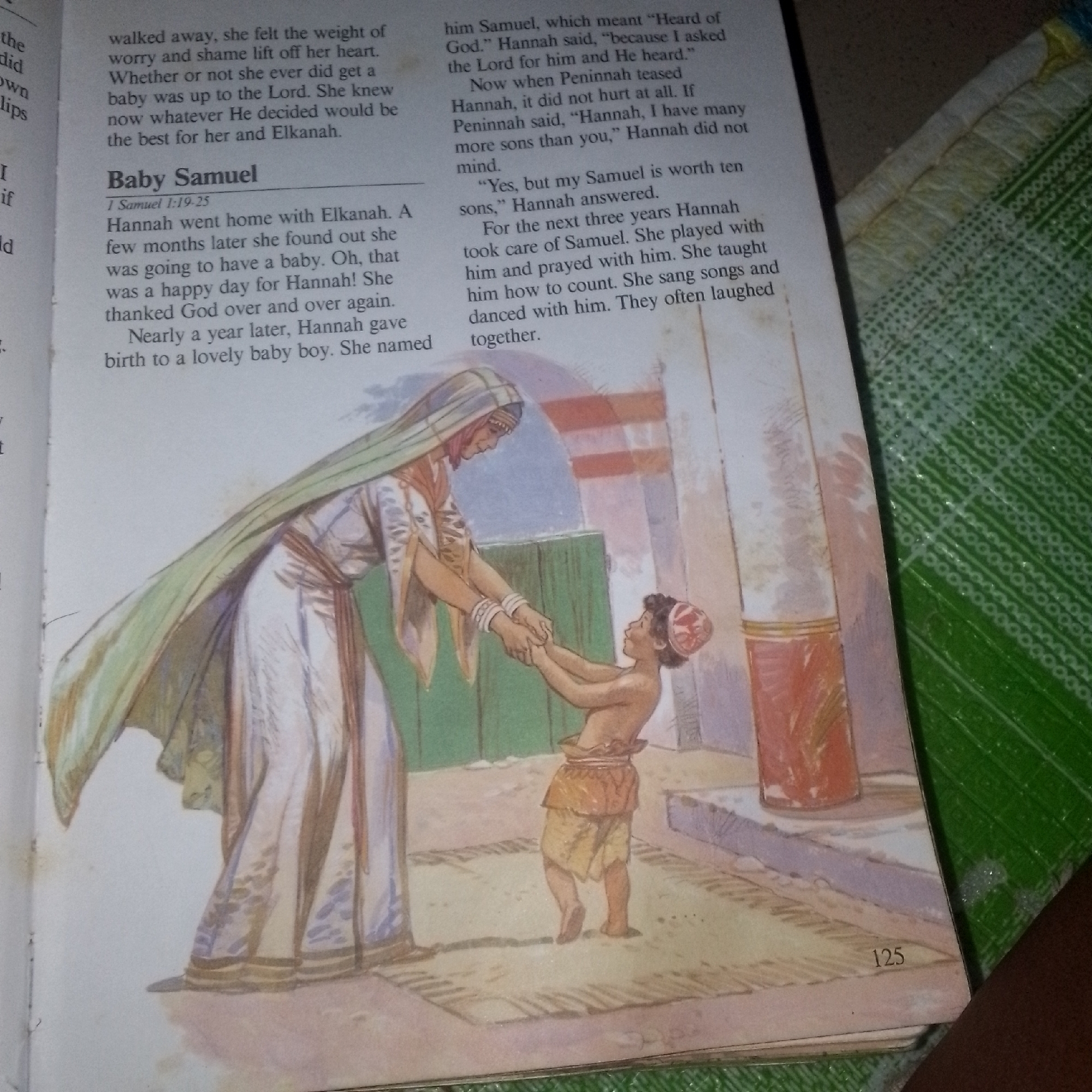 A page from a book displays text and an illustration. The text on the left discusses a woman named Hannah and her desire for a child. The text on the right continues the story of Hannah, detailing her pregnancy, the birth of her son Samuel, and the meaning of his name. It also describes interactions with another woman, Peninnah, and Hannah's upbringing of Samuel.
The illustration depicts a woman in a long, flowing garment and a head covering holding the hands of a young boy. The woman is standing and looking down at the boy, who is facing her and smiling. The boy is wearing a simple skirt. They are standing on a patterned rug in what appears to be an interior setting with architectural elements and green walls in the background. The page number "125" is visible at the bottom right of the illustration.