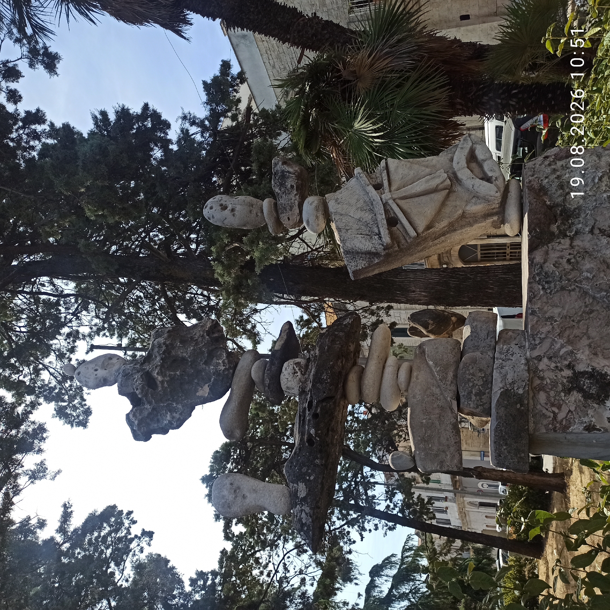 The image depicts an outdoor scene in Ruvo di Puglia, Italy, featuring two large, abstract stone sculptures. The sculptures are composed of stacked and balanced rocks, some of which appear to be carved with rudimentary shapes, including faces or figures with holes. The sculpture on the left has a horizontal stone slab balanced on stacked rocks, with a carved stone object resembling a head on top. The sculpture on the right is more substantial, with a carved element that suggests a face or mask, topped with balanced stones.
The sculptures are situated in a park-like setting with mature trees, including what appears to be a cypress tree and a palm tree, providing shade. In the background, a light-colored building with arched windows and a stone facade is visible, suggesting an urban or semi-urban environment. Vehicles are parked in the background, and there are some leafy green plants in the foreground. The sky is bright and clear, indicating a sunny day. A timestamp "19.08.2026 10:51" is overlaid on the bottom right of the image, suggesting the date and time the photograph was taken. There are no people visible in the scene.