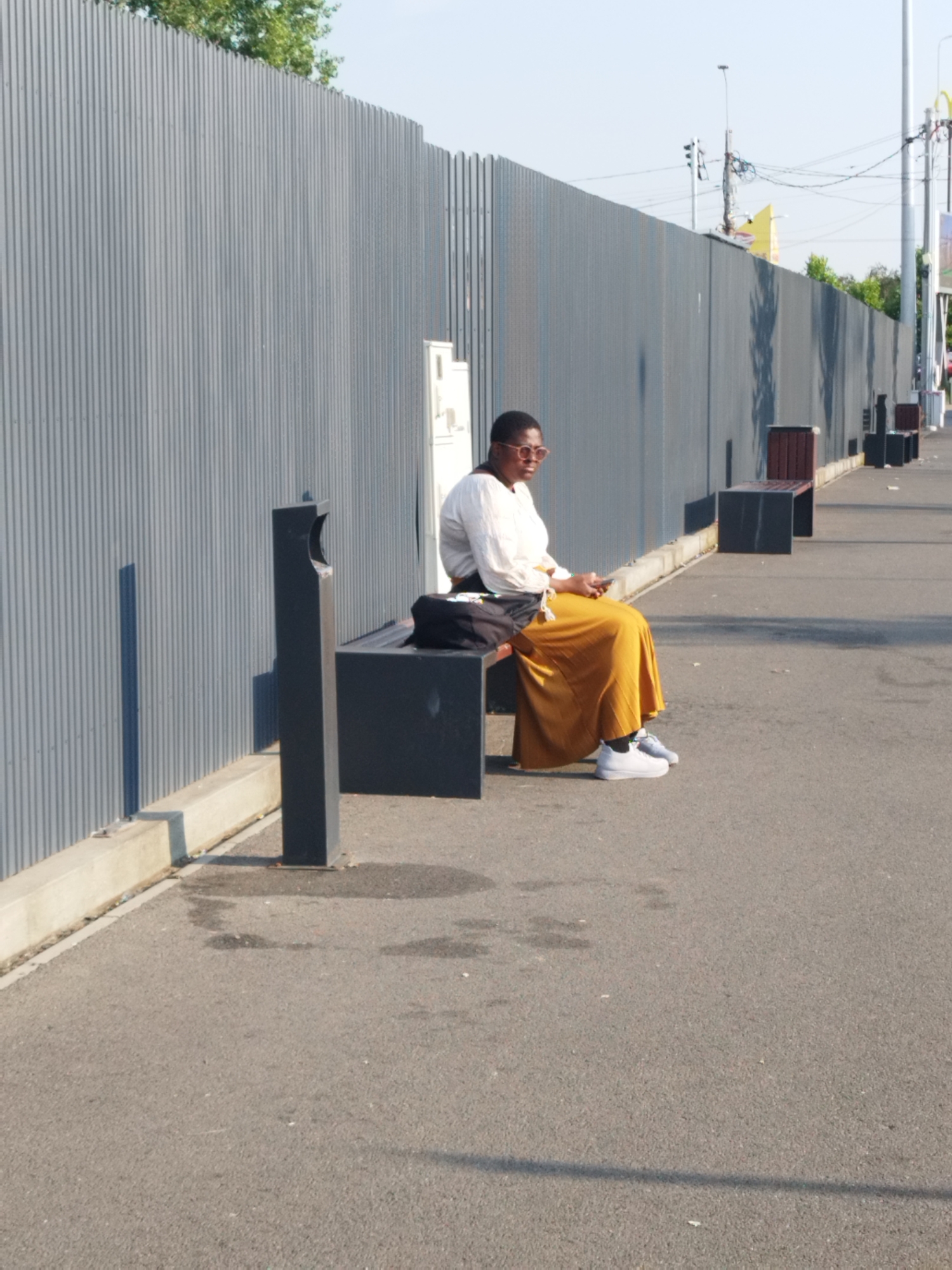In Bucharest, Romania, a woman is seated on a dark-colored bench in front of a corrugated metal fence. She wears a white, long-sleeved top and a flowing, mustard-yellow skirt. Her feet are clad in white sneakers. She is looking down at her phone, which she holds in her hands. A black bag is placed on the bench beside her. To the left of the bench, a black, modern-style trash receptacle with a curved opening stands on the pavement. The scene appears to be outdoors, possibly on a sidewalk or promenade, with an asphalt surface. The lighting suggests it is daytime, with sunlight casting shadows. The fence extends horizontally across the background, and the sky is visible above.