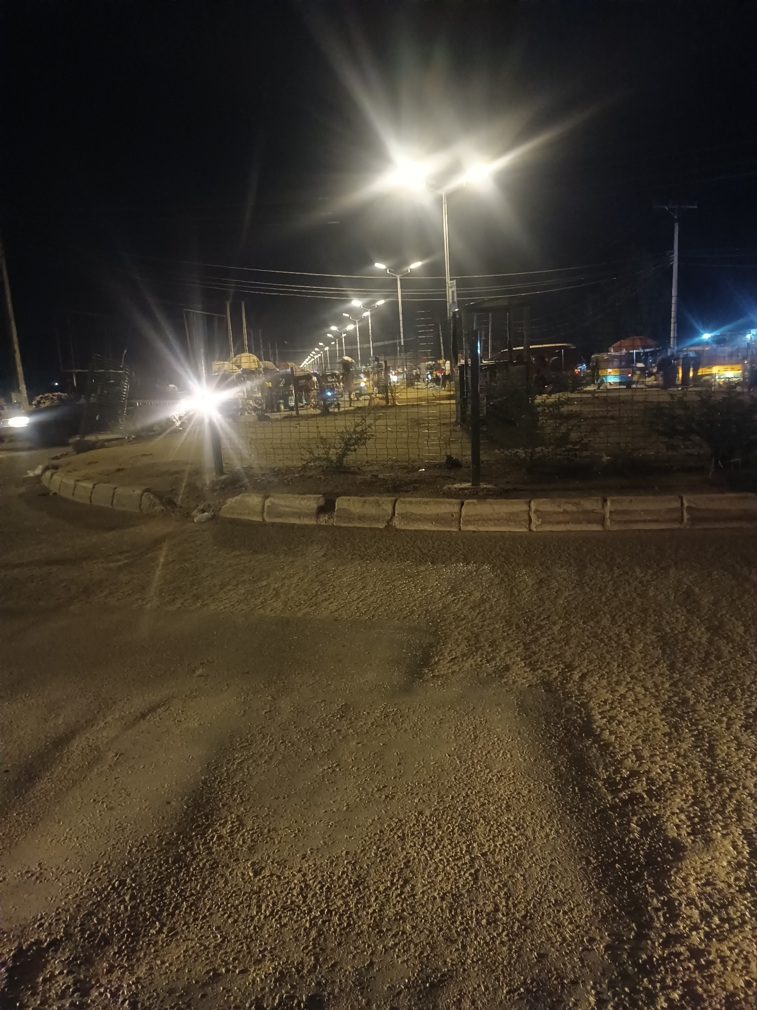 The image shows a street scene at night in Bukuru, Nigeria. The road surface is rough and appears to be covered in loose material. A raised curb separates the road from a fenced-off area with some vegetation. A line of streetlights illuminates the area, creating bright bursts of light. In the background, there are people gathered around what appear to be food stalls or shops. There is vehicular traffic, including at least one vehicle with headlights on. Power lines stretch across the sky above.