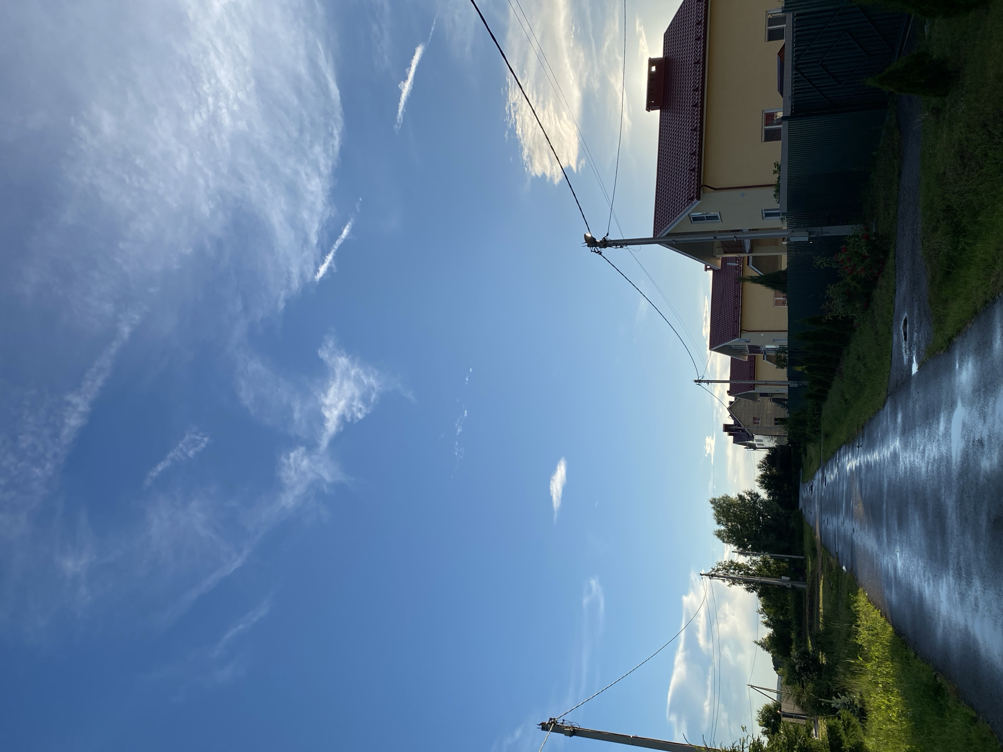 An eye-level, wide shot captures a wet asphalt road curving through a residential area. The road, reflecting the sky, is lined with green grass and trees on the left, and houses with red roofs and yellow walls on the right. Power lines stretch across the frame, supported by utility poles. The sky is a bright blue, dotted with wispy white clouds, suggesting fair weather. No people are visible in the scene. The overall impression is a peaceful, suburban setting.