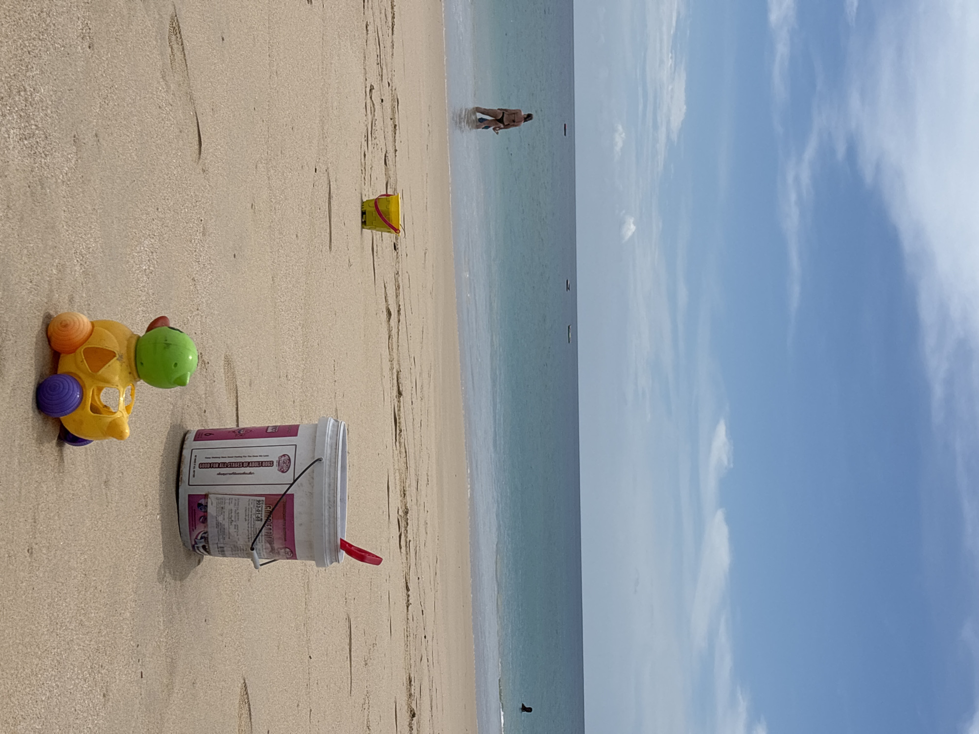 The image shows a beach scene in Choeng Thale, Thailand. The beach is sandy with various objects scattered on the sand including a child's plastic toy car, a white bucket, and a yellow bucket. In the background, there are people in the water. The sky is blue with white clouds.