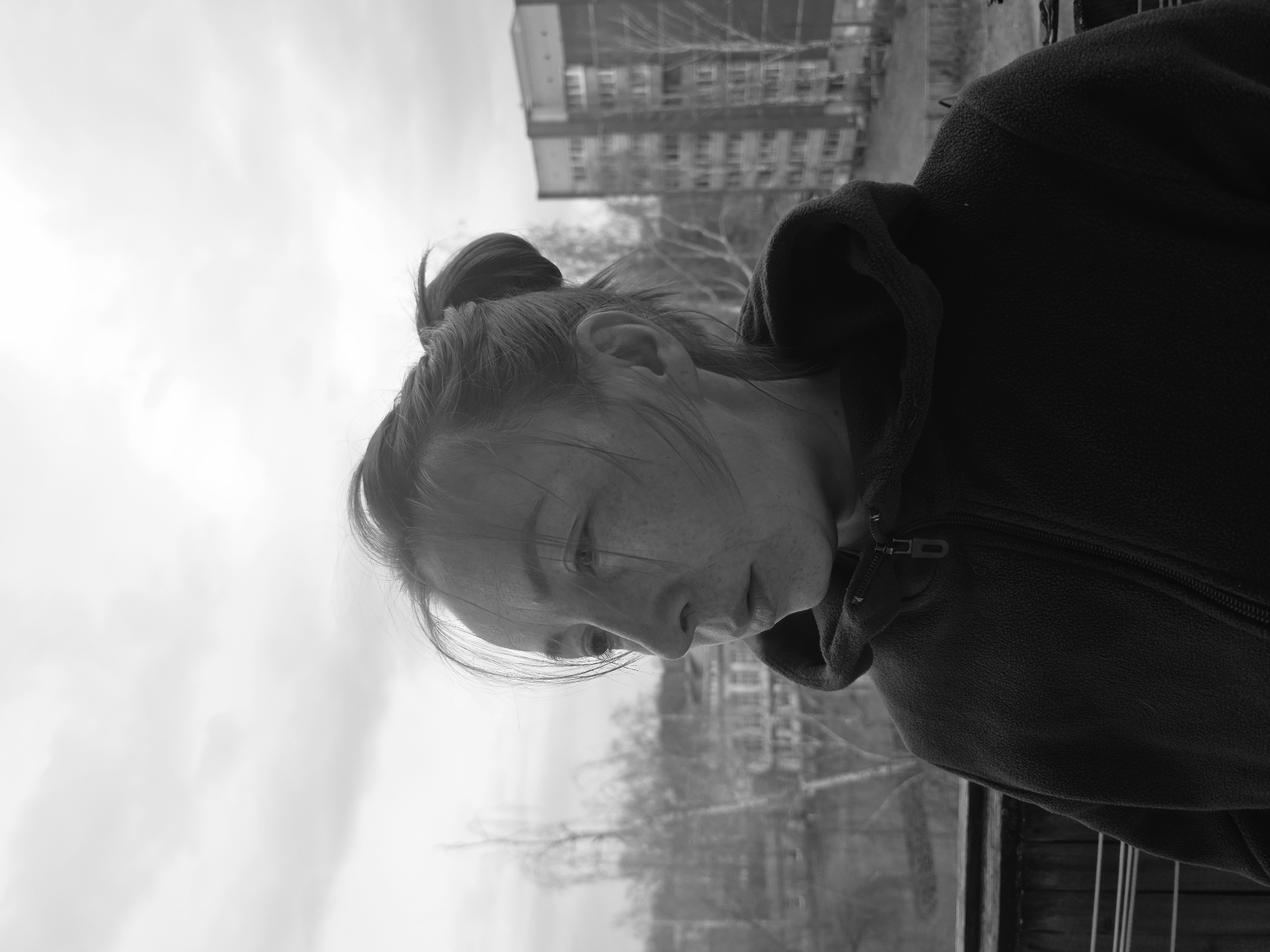 This black and white, vertically oriented photograph captures a young person looking downwards, set against an outdoor, urban backdrop in Chaikovsky, Russia. The image is rotated counter-clockwise, with the person's head on the left and their shoulders extending to the right.
The primary subject is a person, likely a young woman, with her hair pulled back into a bun, and some wisps falling around her forehead and ears. She has visible freckles across her nose and cheeks. Her gaze is directed downwards, suggesting contemplation or observation of something below the frame. She is wearing a dark, possibly fleece, hooded jacket with a visible zipper pull on the right side of the frame. The fabric texture of the jacket is discernable.
The background is softly blurred, indicating a shallow depth of field focusing on the person. A multi-story building, possibly residential or commercial, with scaffolding partially covering its facade, is visible in the upper-right background. Bare trees are also discernible in the middle ground, suggesting a cooler season. The sky is bright and overcast, occupying a significant portion of the upper-left of the frame. A dark railing or fence runs along the bottom-right edge, hinting that the person might be leaning over a balcony or barrier.
The photograph conveys a quiet, perhaps introspective, mood. There is no visible text in the image.