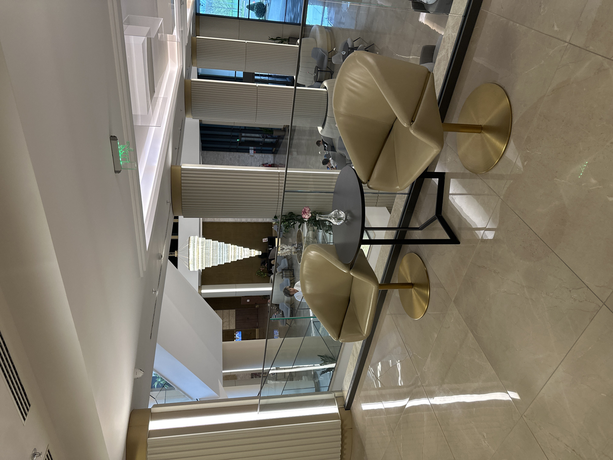 The image captures an upscale, modern interior space, likely a lobby or lounge, in Kazanlak, Bulgaria. The perspective is from an elevated vantage point, looking down into a lower level through a clear glass railing.
In the foreground, two contemporary armchairs are prominently featured. They have light beige or cream-colored upholstery with a distinctive geometric fold pattern and are supported by sleek, polished gold-colored circular bases. Between these chairs is a small, dark grey round table, on which a clear glass vase holds a single pink flower.
The background reveals the lower level of the building, accessible via a large open space. This area is furnished with various seating arrangements, including curved sofas and individual chairs, several of which are occupied by people who appear to be waiting or socializing. Architectural elements include tall, vertically ribbed panels lining the walls, and a grand, multi-tiered chandelier composed of numerous clear crystalline segments hangs from the ceiling. Large windows in the background allow natural light to illuminate the space, and glimpses of outdoor greenery are visible.
The flooring throughout is made of polished, light-colored marble or ceramic tiles, which reflect the abundant lighting. The ceiling features a modern design with integrated strip lighting, recessed fixtures, and air vents. A green exit sign, depicting a running person symbol, is mounted on a wall in the upper-left of the image. The overall impression is one of sophistication and brightness, suggesting a daytime setting.