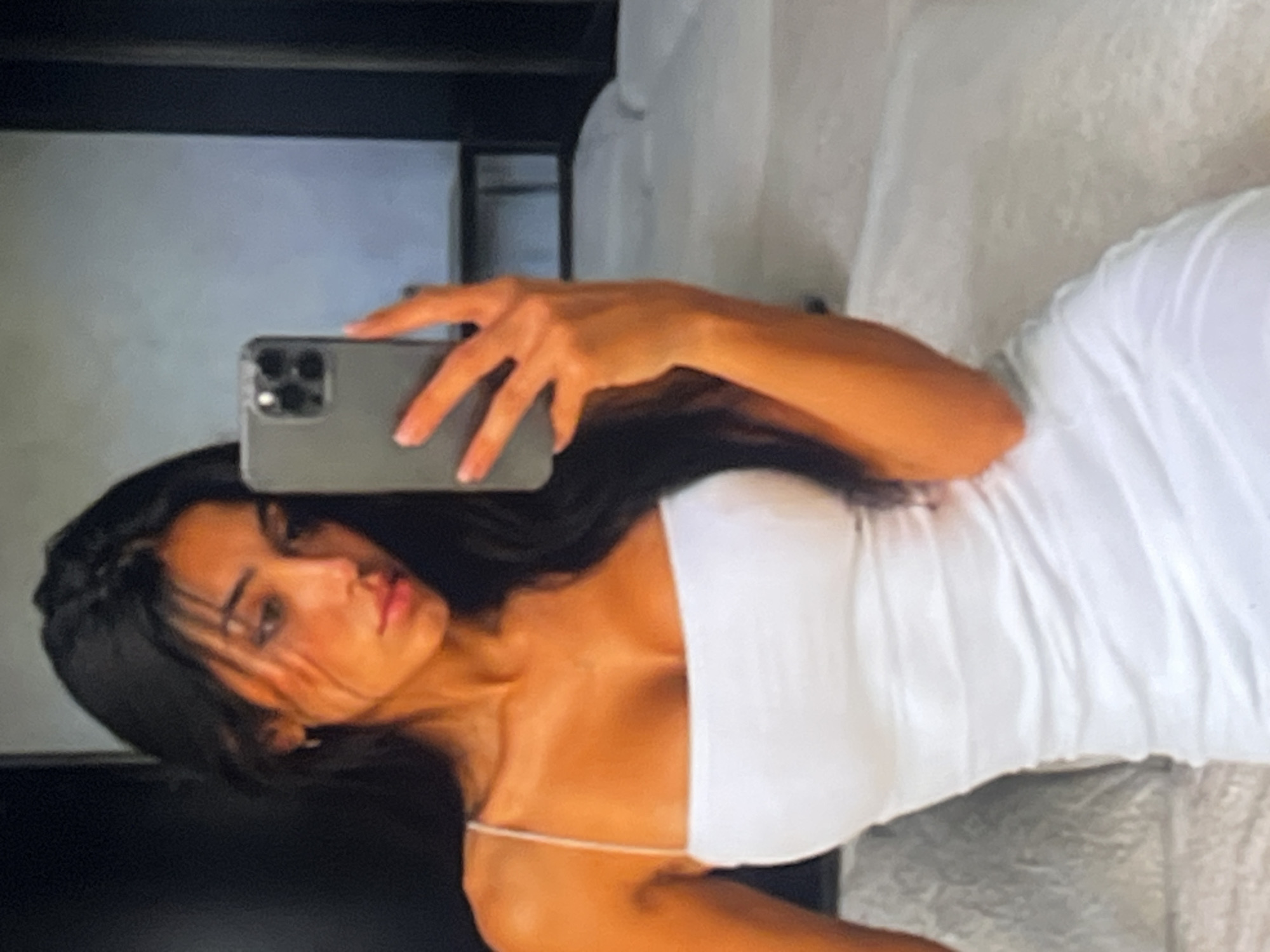 A woman with long, dark hair and a white, strapless dress is captured in a mirror selfie. She is holding a gray smartphone in her right hand, which is positioned in front of her chest. The background shows a bed with white sheets and pillows, suggesting an indoor setting, possibly a bedroom. The lighting is warm, casting a golden hue on the woman's skin.