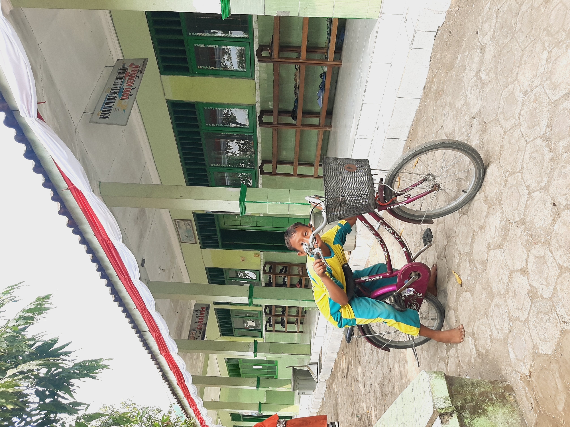 In Sumberjo, Indonesia, a young boy with a bright yellow and blue shirt and teal pants sits on a maroon bicycle in front of a light green building. He is barefoot and looks directly at the camera with a slight smile. The bicycle has a black wire basket on the front and ornate designs on the frame. The building appears to be a school, with a covered walkway supported by white pillars, decorated with draped red and white fabric. Green-barred windows line the building's facade, and wooden shoe racks are visible inside the walkway. The ground is paved with hexagonal concrete tiles. To the left of the boy, a portion of a light green concrete bench is visible, and a woman in an orange hijab and skirt can be seen in the distance. The sky is bright and overcast.
