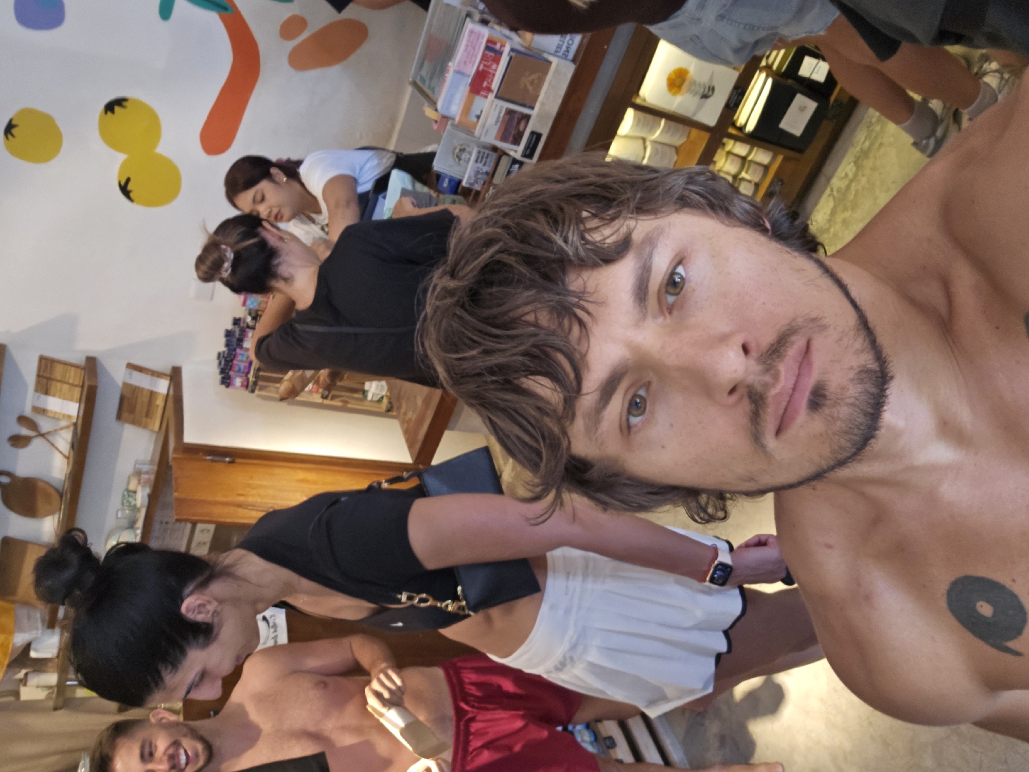A selfie taken in an indoor establishment, potentially a shop in Tibubeneng, Indonesia. A light-skinned man with wavy brown hair and a beard occupies the foreground, shirtless except for a tattoo on his right pectoral. In the midground, there are at least three other people: one fair-skinned person in red shorts and two people interacting with each other. One is wearing a black top and white skirt. The background includes shelves of products, a wall with painted shapes, and an open space with additional people. The lighting appears to be natural and diffused.