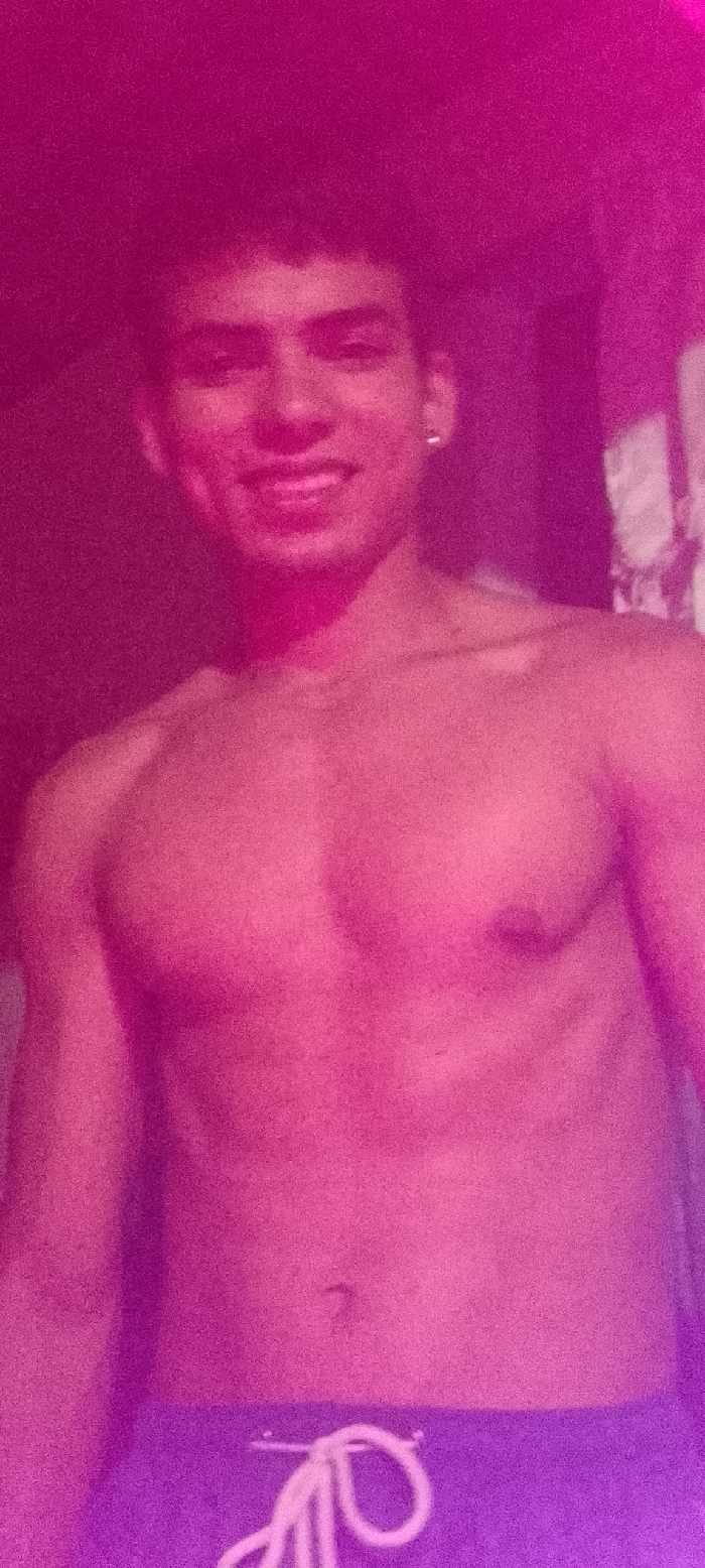 A shirtless young man smiles at the camera. He has short, dark hair, dark eyes, and an earring in his left ear. He wears a blue garment with a white drawstring at the waist. The image has a pink and purple hue, which impacts the skin tone and overall visibility of the subject. The background is dark and indistinct, likely indoors.