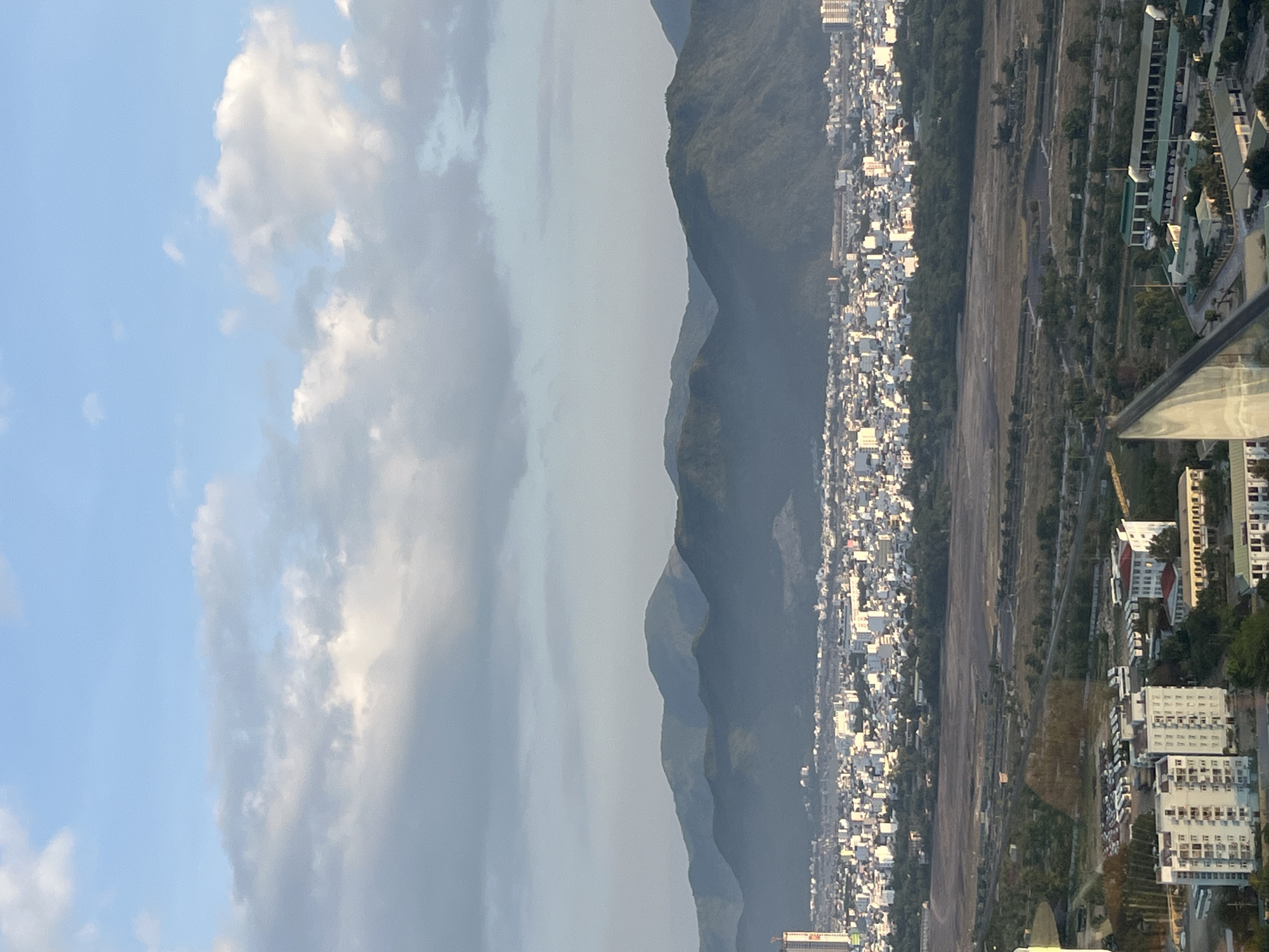 This high-angle panoramic image depicts a vast urban and natural landscape in the city of Nha Trang, Vietnam, viewed under a partially cloudy sky.
The upper third of the image shows a light blue sky with scattered clouds. Some clouds are bright white and illuminated by sunlight, while others are a darker gray, casting shadows or indicating greater thickness.
Below the sky, a distant range of dark green, rounded mountains stretches across the horizon, appearing hazy. At the base of these mountains, an extensive cityscape unfolds, characterized by a dense cluster of predominantly white and light-colored buildings with varying roof tones. Sunlight highlights the upper portions of these structures. A single tall, slender building, potentially under construction, with a red and white top, is prominently visible on the far left side of the urban sprawl.
In the middle ground, separating the distant city from the immediate foreground, lies a large, relatively undeveloped area of brownish-gray earth, possibly fields or construction sites. This area is traversed by a network of visible roads and interspersed with patches of green vegetation, including small trees and bushes.
The immediate foreground, occupying the lower third of the image, displays more detailed architectural and environmental elements. On the left, a multi-story residential or commercial building with numerous windows and balconies is visible. Adjacent to it, a paved lot contains a high density of parked white vehicles, likely vans or small buses. Further right, another group of buildings, some with green-colored roofs, are present. A distinctive architectural element, possibly a triangular glass facade reflecting light, occupies the lower right edge of the frame, suggesting the vantage point is from inside a modern building looking out. No people or specific human activities are discernibly visible within the scene.