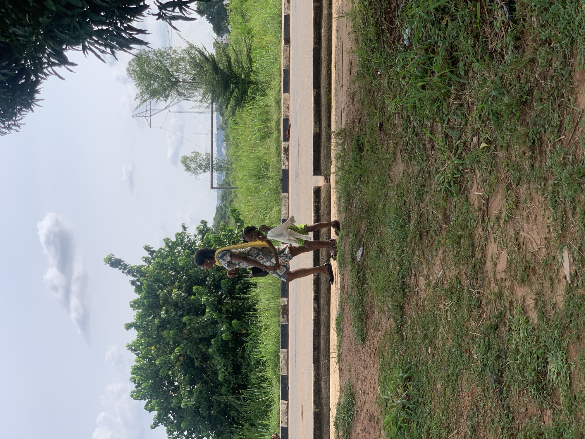 A woman and a child walk along a paved road in an outdoor, urban environment. The woman is on the left, wearing a patterned dress and a yellow headscarf, and carries a dark bag. The child, to her right and slightly behind, wears a green outfit and carries a white cloth. They walk side-by-side on the road's edge, near a curb with black and white markings. The background features lush green vegetation, tall grass, trees, and distant power lines under a cloudy sky. The foreground is a patch of reddish-brown dirt and grass.