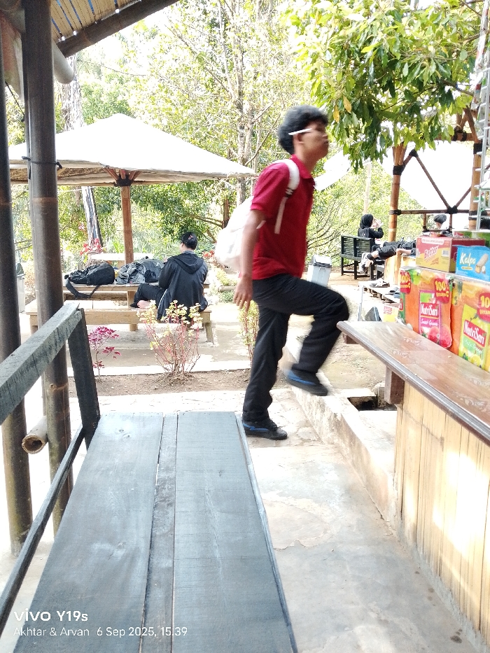 The image captures an outdoor scene in Sukadana, Indonesia, on September 6, 2025, at 15:39.
In the immediate foreground on the left, a dark wooden plank bench is visible, alongside a bamboo support column. A male individual occupies the mid-ground, captured in motion, stepping onto a raised concrete platform with his right foot. He wears a red short-sleeved polo shirt, dark trousers, and dark athletic shoes with blue accents. A white backpack is worn on his shoulders, and a white object is positioned behind his right ear. His head is turned slightly to his left, and he wears glasses.
To the right of the moving individual, a wooden display counter is stocked with various packaged items, including multiple orange boxes labeled "NutriSari," a blue box labeled "Kalpa," and a yellow box labeled "Batter."
Further in the background on the left, under a large white umbrella, a person is seated on a wooden bench, facing away from the camera. This individual wears a dark long-sleeved top and dark trousers. Several dark bags are resting on the bench next to them. Small plants with reddish foliage are visible in the ground strip separating the foreground bench and the seated person.
The background consists of dense green foliage from numerous trees, indicative of a natural outdoor setting under bright daylight. In the far background, on the right, additional individuals are visible, seated on a bench beneath a bamboo-supported shelter. One of these individuals is wearing a head covering. The ground throughout the scene is a combination of concrete pathways and exposed soil.