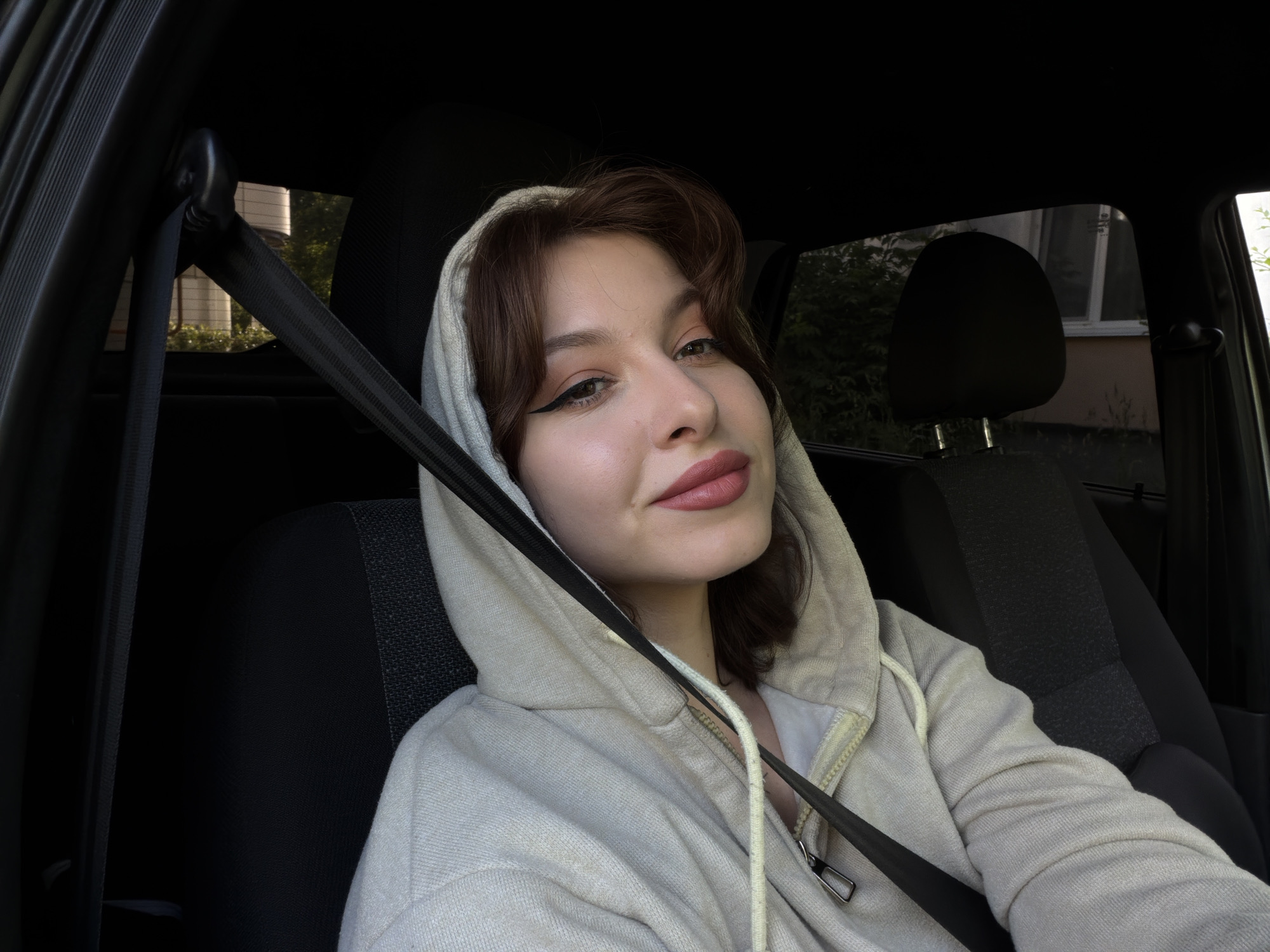 The image is a selfie taken by a young woman sitting in the passenger seat of a car. She is wearing a light-colored hooded sweatshirt and has her seatbelt fastened across her chest. Her hair is brown and styled loosely around her face. She is looking towards the camera with a slight smile.
The setting appears to be daytime, with natural light filtering into the car. Through the car windows, trees and a building are visible, suggesting an outdoor location, possibly residential or suburban. The interior of the car is visible, including the black car seat, seatbelt mechanism, and part of the dashboard.
The woman is the primary subject of the image. She has makeup on, including eyeliner and lipstick. Her expression is relaxed and perhaps a little playful. There are no visible signs of text in the image. The overall mood of the image is casual and personal.