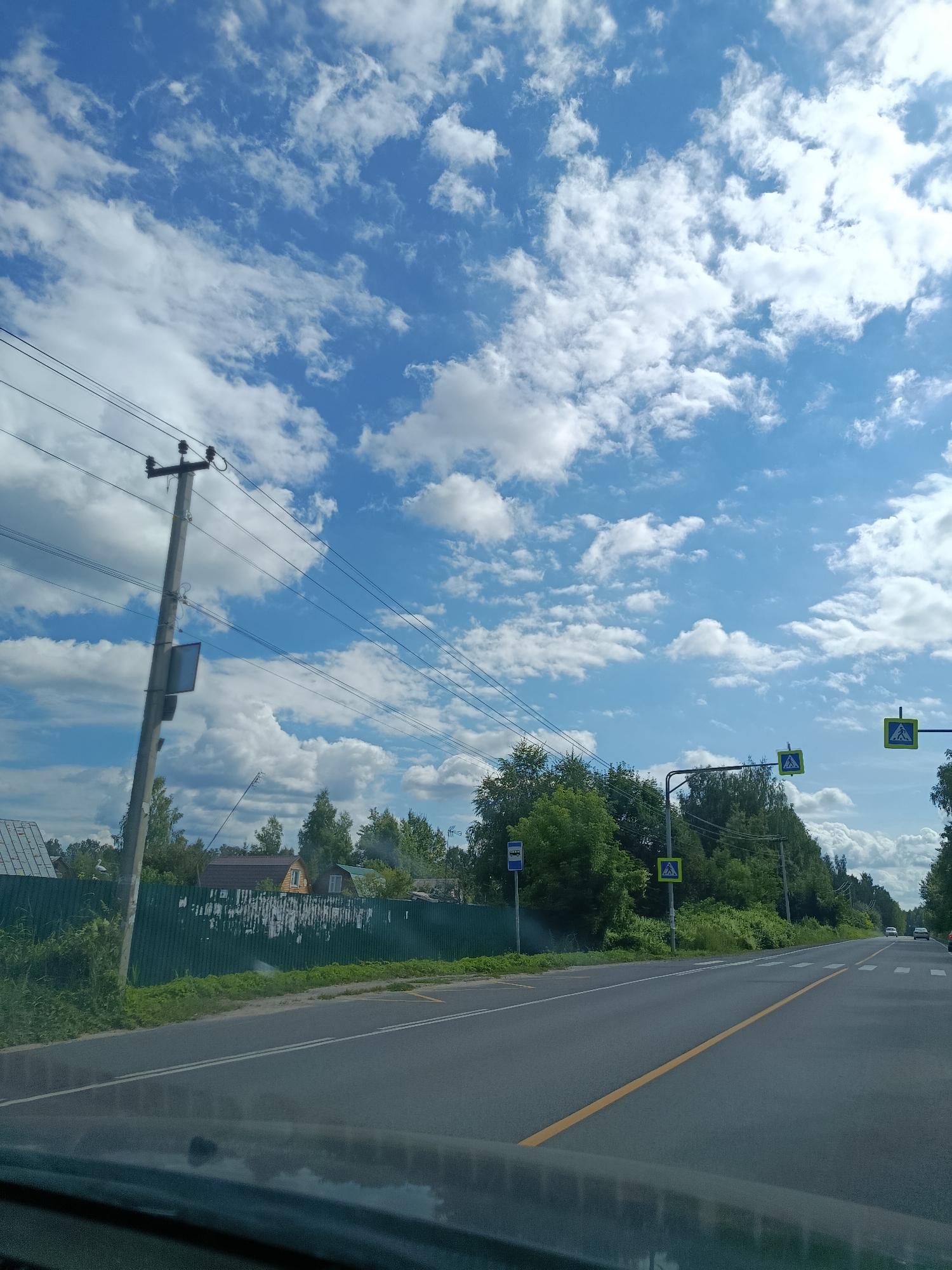 The image depicts a view from a vehicle driving on a two-lane asphalt road. The road has a yellow centerline and white lane markers. On the right side of the road, there are pedestrian crossing signs with yellow triangular backgrounds and white pedestrian symbols. A bus stop sign is visible on the left side of the road, along with a power pole with multiple wires. Behind the road on the left, there is a green metal fence, some overgrown vegetation, and residential buildings. On the right side, there is a dense line of trees. The sky is bright blue with scattered white clouds. There are two cars visible in the distance on the road. The scene appears to be in a rural or suburban area, possibly on the outskirts of 1-Go Maya, Russia, given the presence of residential structures and a road that transitions from a more built-up area to a more natural landscape. No people are directly visible in the image.