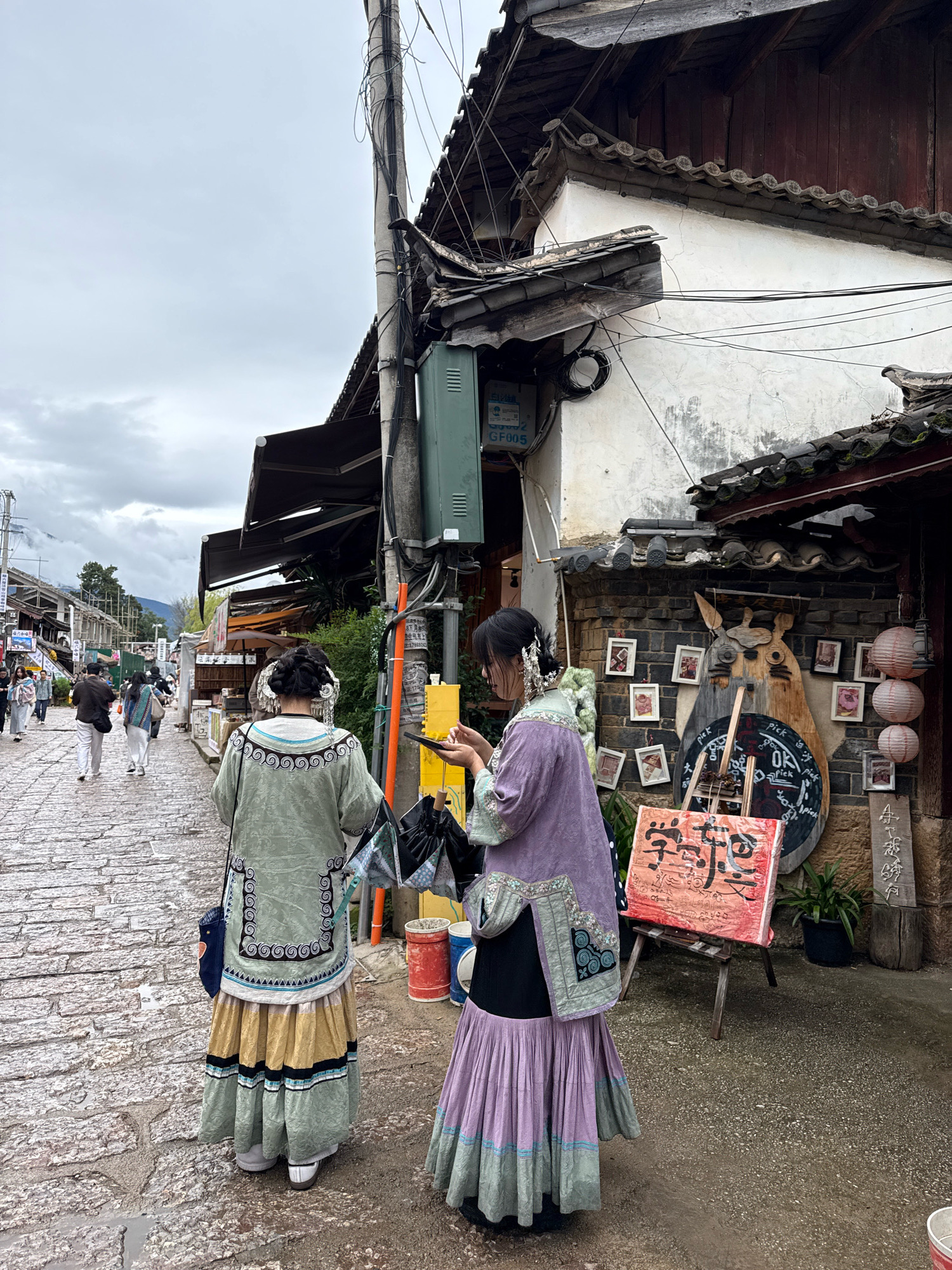 Two women in traditional ethnic clothing are walking down a cobblestone street in Yulong Nakhi Autonomous County, China. The woman on the left wears a green tunic with intricate embroidery and a multi-layered skirt in shades of yellow and green. The woman on the right wears a purple tunic with similar embroidery over a black skirt and a layered purple skirt. They are both facing away from the camera. Behind them, a street is lined with traditional buildings with tiled roofs. There are several shops or stalls along the street, one of which displays a wooden carving of a character that resembles Totoro, surrounded by framed pictures. A red sign with Chinese calligraphy is propped on an easel in front of the stall. The sky is overcast.