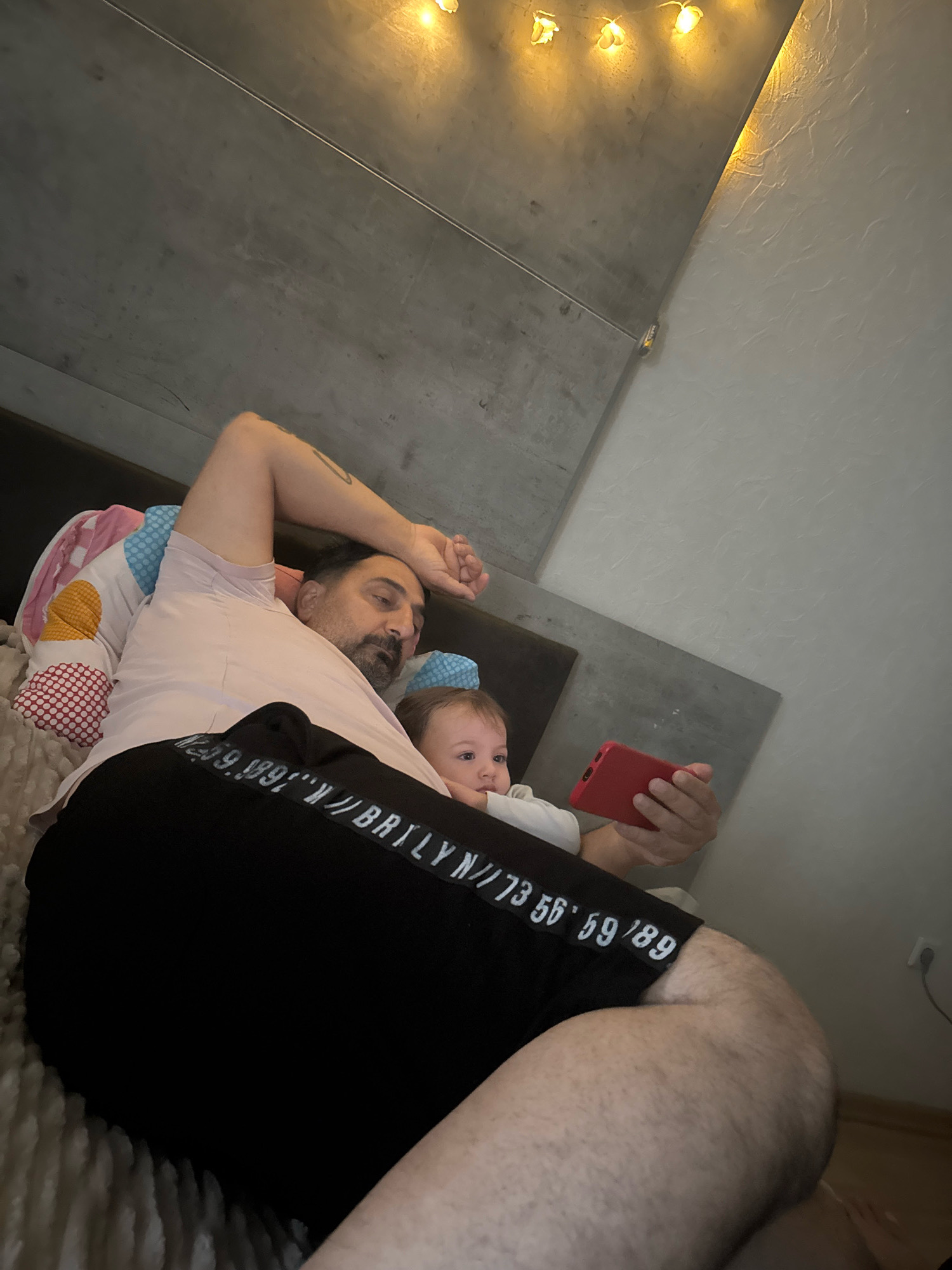 In a bedroom in Überherrn, Germany, a man and a baby lie in bed. The man, with dark hair and a beard, is wearing a light pink t-shirt and black shorts. He is holding a red smartphone in his right hand, and his left arm is raised above his head, resting on a pillow. The baby, with light brown hair, is leaning against the man's chest, looking at the smartphone. The bed has pillows and a fluffy blanket. The background shows a textured wall and a dark, possibly concrete-like, ceiling adorned with string lights.